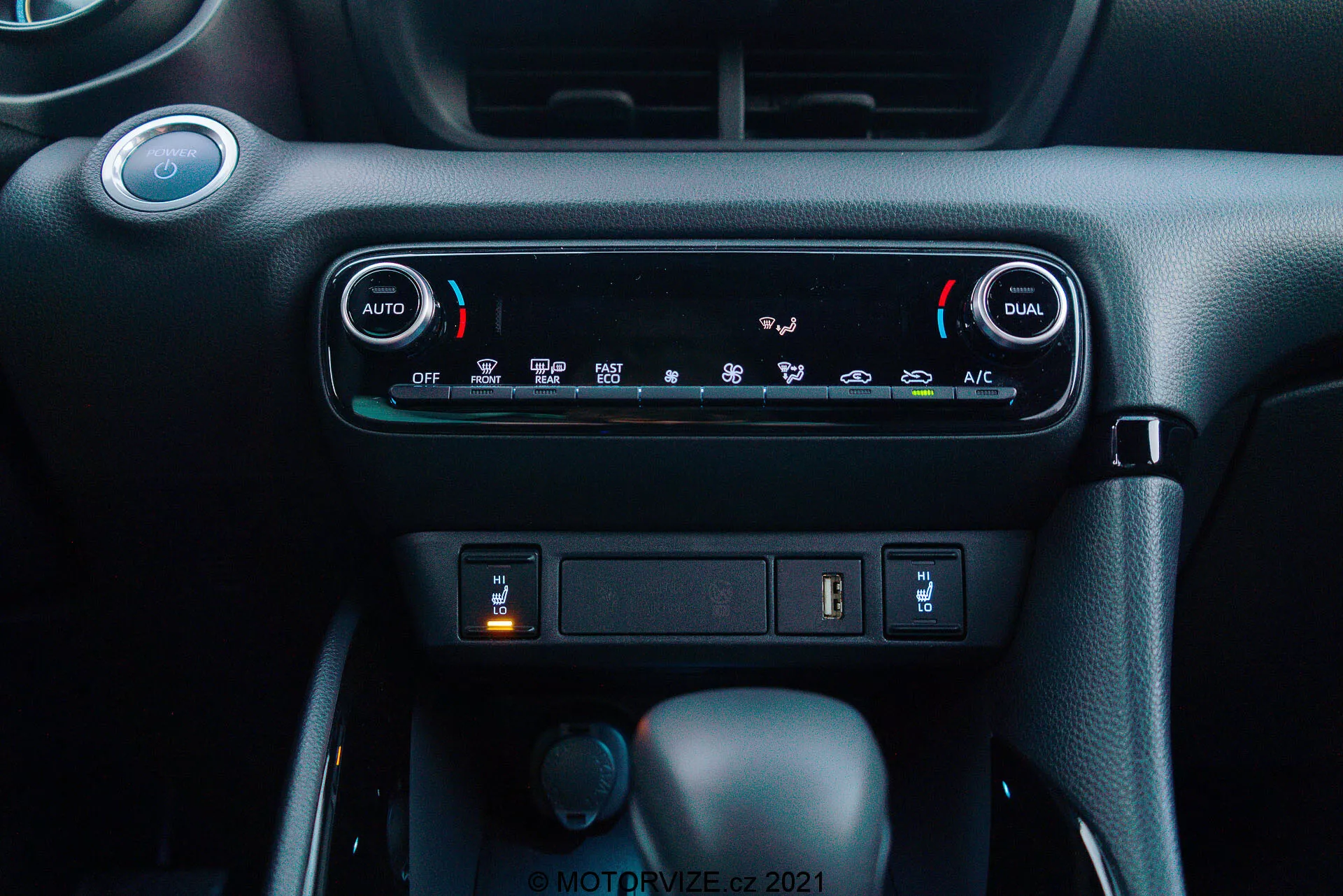 Vista aproximada do painel do Toyota Yaris 2020: A imagem destaca o sistema de controle climático de zona dupla automática, com botões 'AUTO' e 'DUAL' para ajustes de temperatura independentes para o motorista e passageiro. Abaixo dos controles de climatização, há um compartimento com uma entrada USB e uma possível tomada de 12V. Mais abaixo, três botões são visíveis: o central é a alavanca de câmbio no modo 'Park', indicando uma transmissão automática, um botão rotativo menor à esquerda provavelmente para seleção de modo de condução, e um botão à direita que pode ser um freio de estacionamento eletrônico. O material do painel tem um esquema de cores escuras com acabamento texturizado premium, criando um ambiente de cabine moderno e fácil de usar.