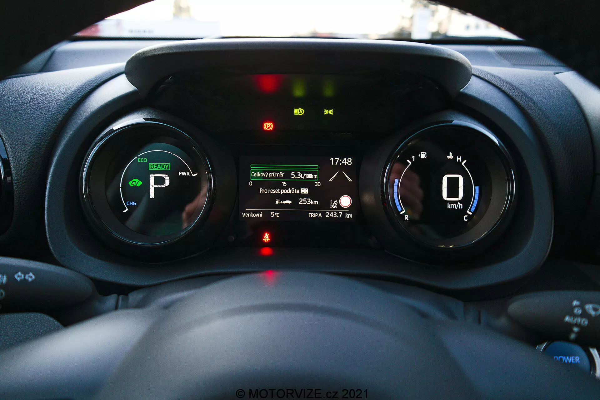 Vista interior do painel do Toyota Yaris 2020 focando no painel de instrumentos, tirada da perspectiva do motorista mostrando medidores digitais, indicadores de veículo elétrico ou híbrido, e uma tela de múltiplas informações com recursos como distância da viagem, temperatura externa e faixa da bateria.