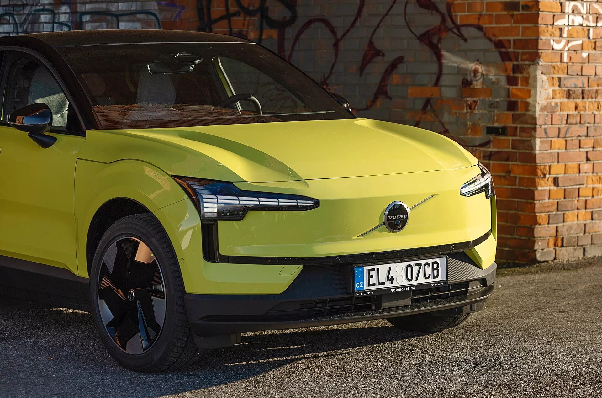Carro elétrico Volvo EX30 verde lima de 2024 estacionado em frente a um muro de tijolos com graffiti, exibindo design aerodinâmico, faróis LED modernos e ausência de grade frontal