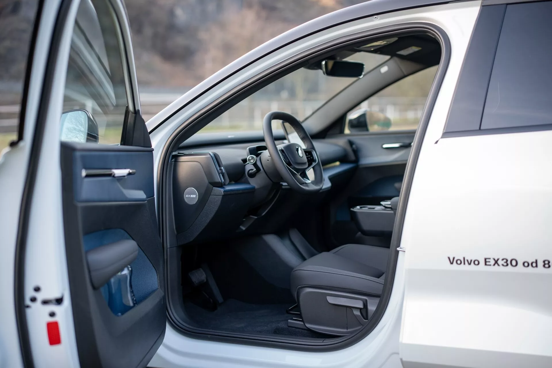 Vista interior de um Volvo EX30 2024 branco, mostrando o assento do motorista e área do cockpit com a porta da frente esquerda aberta apresentando controles elétricos, e um vislumbre do volante e parte do painel