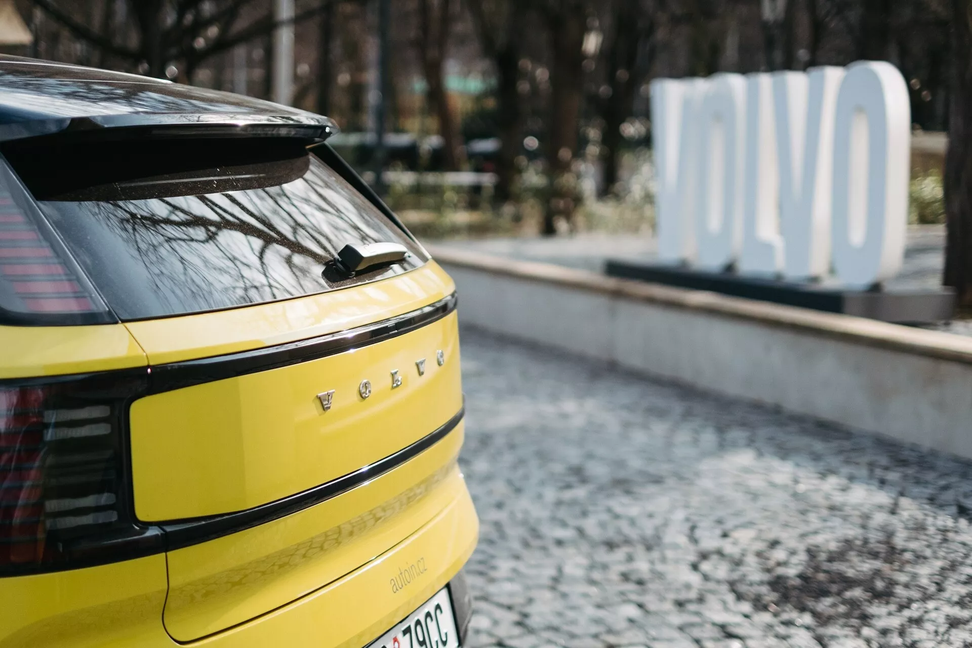 Close-up da seção traseira de um Volvo EX30 2024 amarelo brilhante, apresentando luzes traseiras LED horizontais, potencial câmera ou sensores traseiros, limpador de para-brisa traseiro único, emblema de veículo elétrico ou híbrido e antena em forma de barbatana de tubarão, estacionado em superfície de paralelepípedos com um sinal 'OLVO' desfocado no fundo