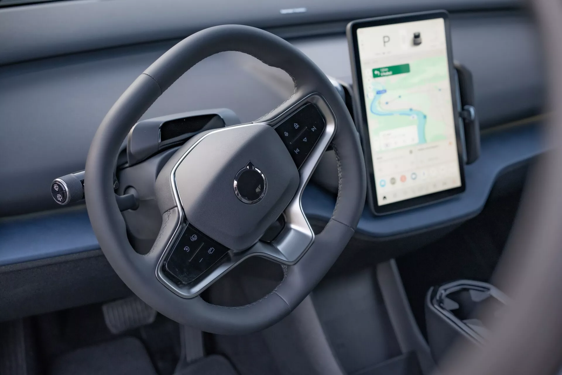 Vista interior do Volvo EX30 2024 destacando a área de controle do motorista, com um volante moderno de fundo plano e um grande display touchscreen vertical para navegação e mídia no painel