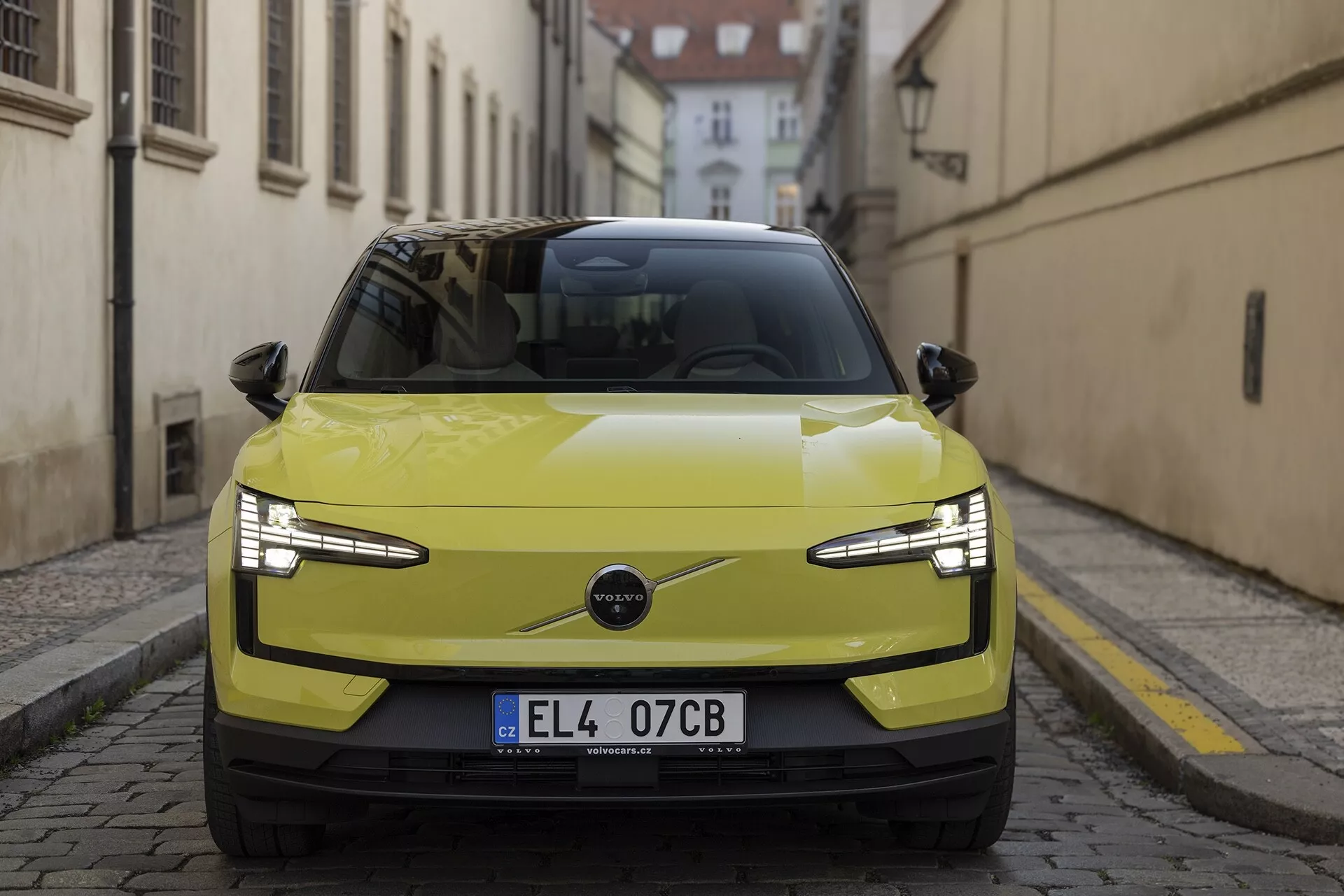 Vista frontal do Volvo EX30 2024 em amarelo lima, destacando seus faróis LED elegantes, grade minimalista indicativa de um tren de força elétrico e recursos tecnológicos avançados como sensores de estacionamento e possível ADAS, contra uma rua europeia histórica
