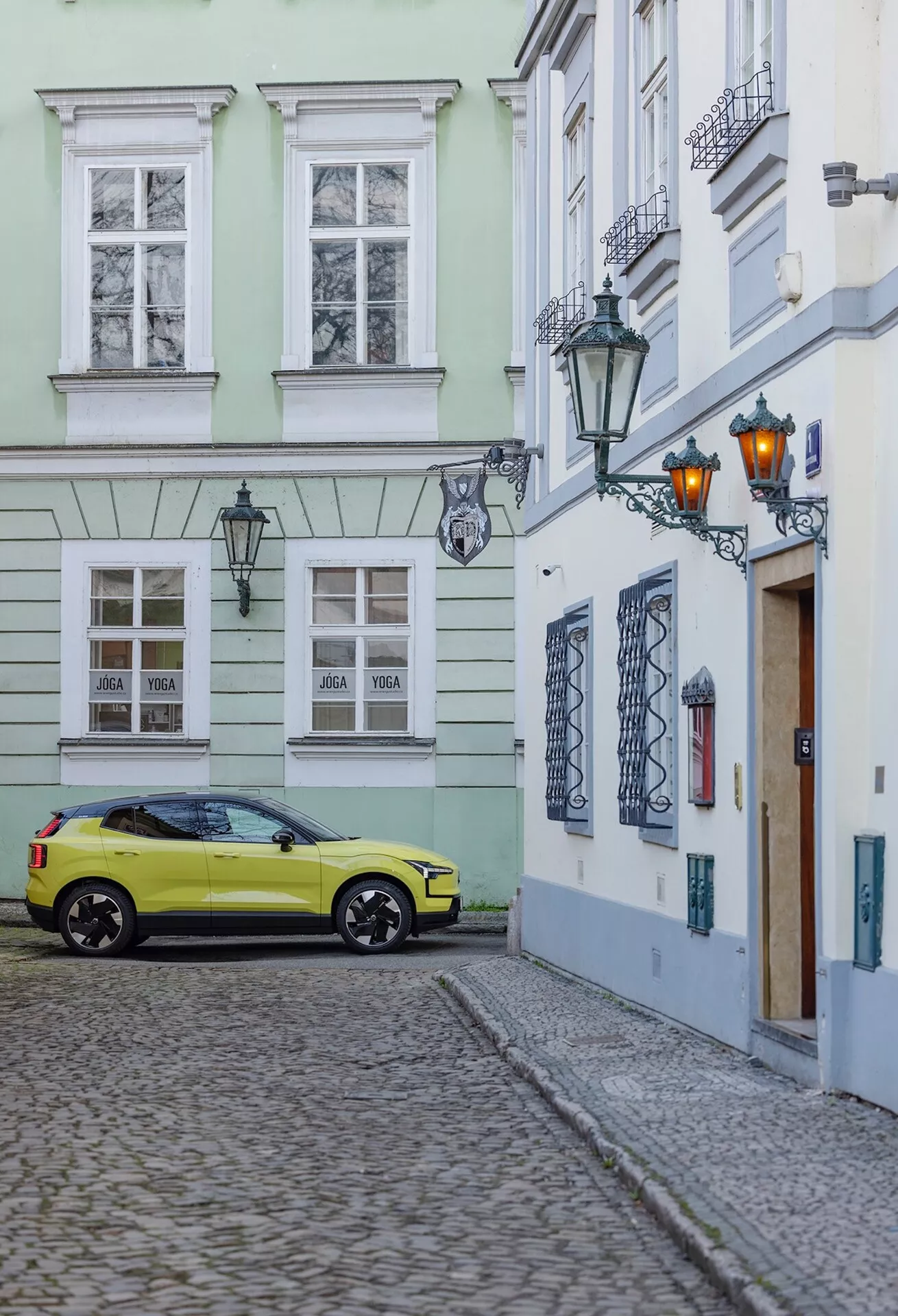Volvo EX30 2024 estacionado em uma rua de paralelepípedos dentro de um cenário de cidade antiga, capturado de um ângulo traseiro de três quartos mostrando o lado direito e traseiro do carro incluindo as rodas, luzes traseiras e detalhes parciais do teto na cor amarela brilhante