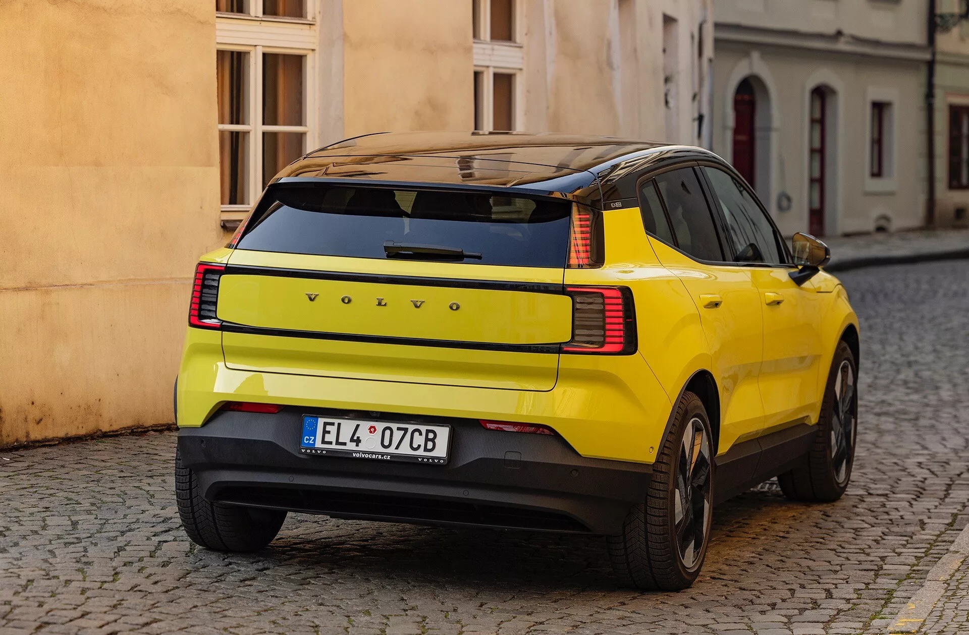 SUV amarelo Volvo EX30 2024 exibindo sua curvatura distinta do painel traseiro, luzes traseiras LED e características de segurança, estacionado em uma rua de paralelepípedos vista de três quartos de trás