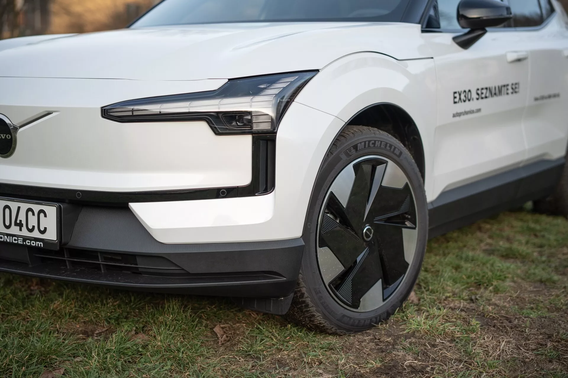 Vista de perto do veículo elétrico Volvo EX30 2024 de uma perspectiva de três quartos, destacando seu farol LED, design aerodinâmico da roda e para-choque dianteiro minimalista sem grade, em branco com acabamentos em preto.