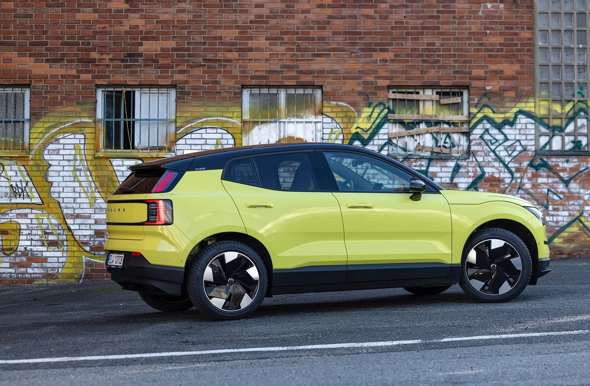Imagem lateral de um SUV Volvo EX30 verde-lima de 2024 estacionado ao lado do meio-fio de uma rua urbana com arte de grafite ao fundo, exibindo suas rodas de liga leve pretas esportivas, luzes traseiras LED e luz de stop alta.