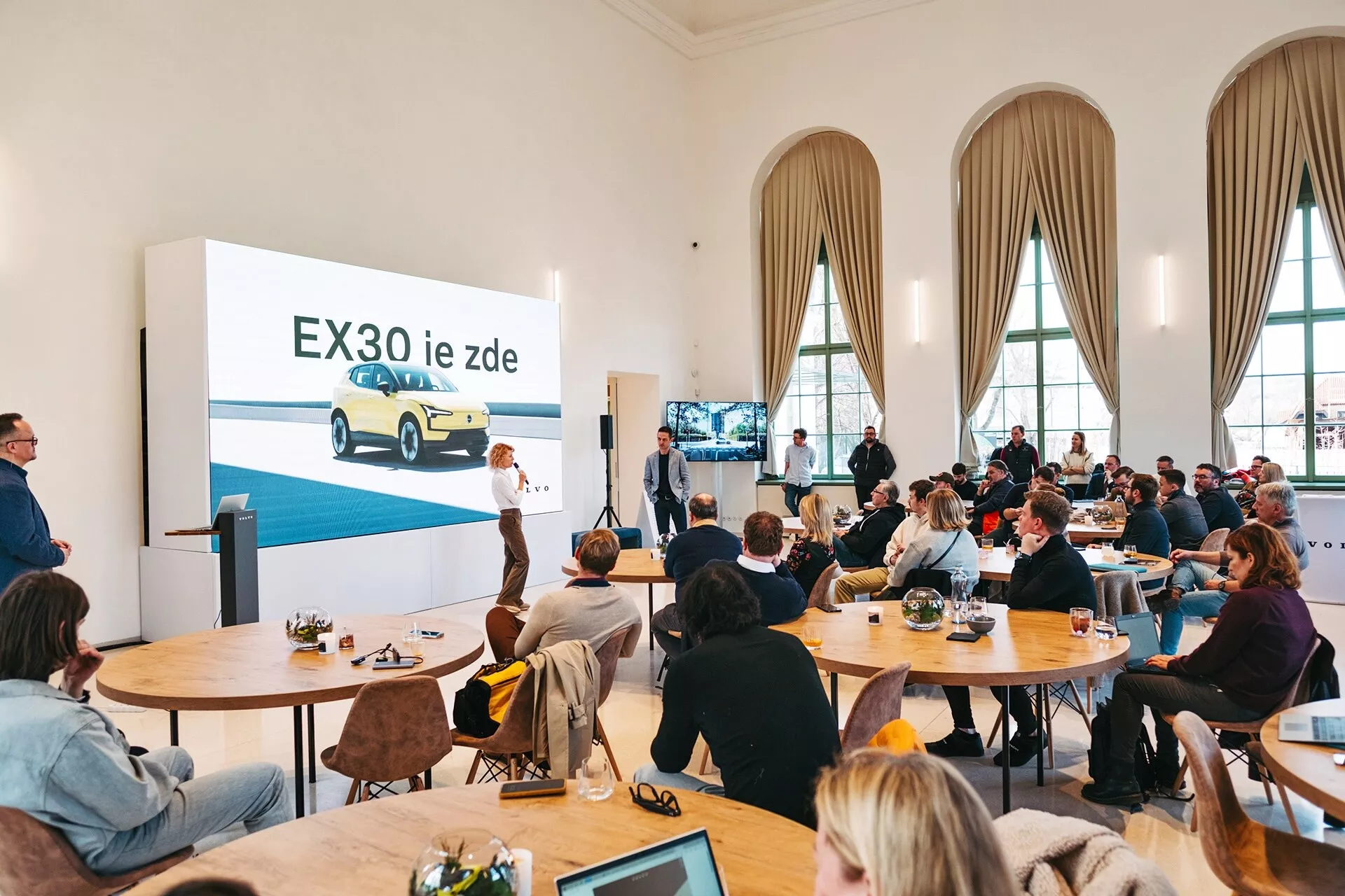 Apresentação do veículo elétrico Volvo EX30 2024 em uma sala espaçosa com tetos altos e grandes janelas arqueadas, apresentando um EX30 amarelo de uma visão frontal de três quartos em uma grande tela