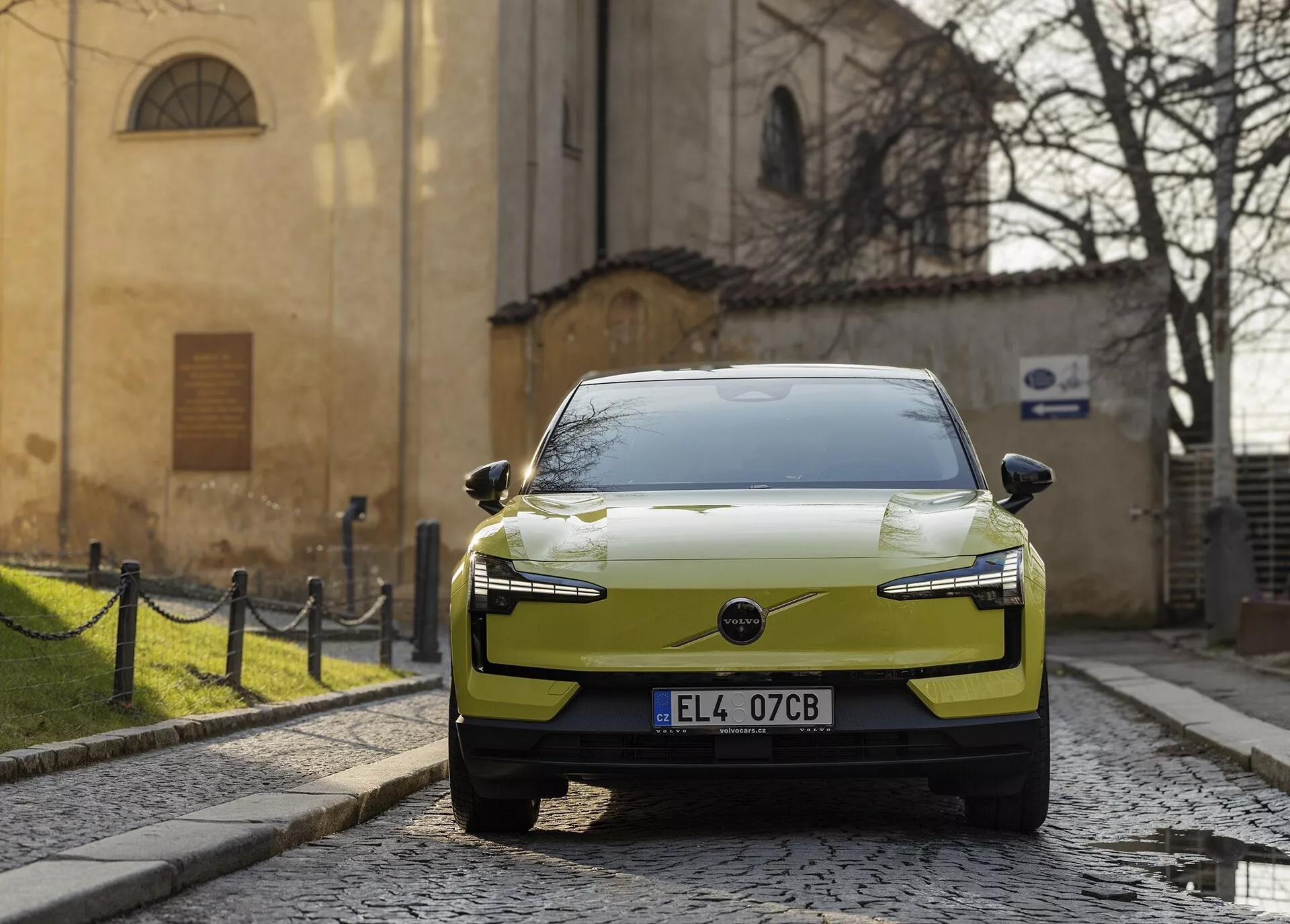 Carro elétrico moderno Volvo EX30 de 2024 na cor chartreuse, estacionado em uma rua de paralelepípedos, visto de uma perspectiva frontal-direita com faróis LED e um design aerodinâmico do capô realçado contra um cenário histórico europeu.
