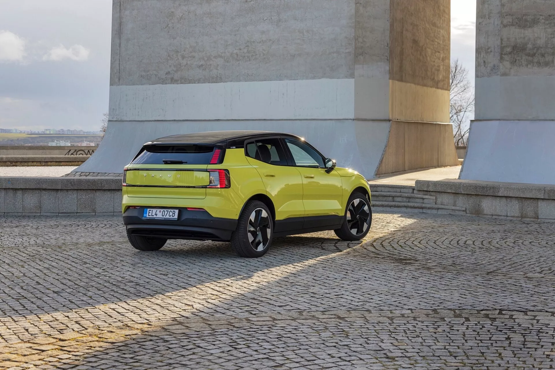 SUV verde-lima Volvo EX30 2024 em vista traseira de três quartos mostrando perfil lateral, lanternas traseiras, porta-malas e rodas de liga preta perto de paredes de pedra em chão de paralelepípedos