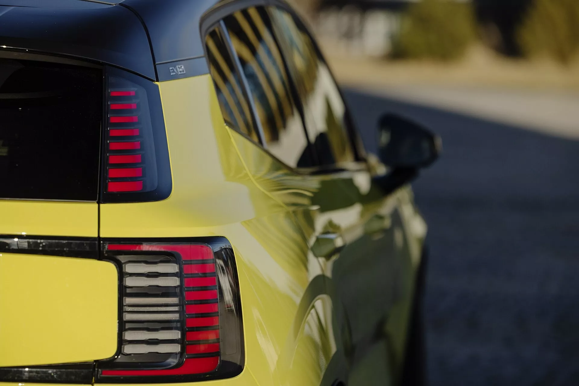 Visão detalhada do quarto traseiro de um Volvo EX30 2024 em amarelo vibrante, destacando o acabamento brilhante, lanternas traseiras LED segmentadas, linhas de design aerodinâmico e sinais de mudança de direção integrados no espelho lateral