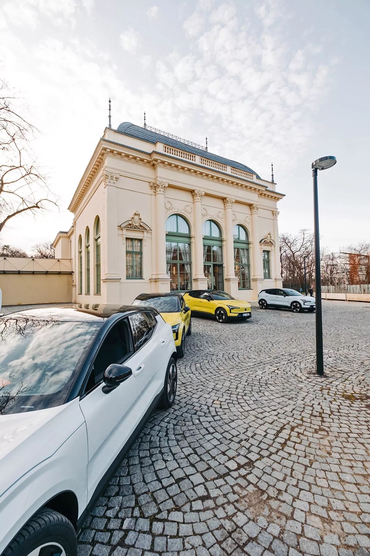 Cena de rua com três carros e um edifício elegante ao fundo, capturada de uma perspectiva de grande ângulo destacando um sedan contemporâneo branco, um carro esportivo amarelo vibrante e um sedan ou hatchback branco à luz do final da tarde