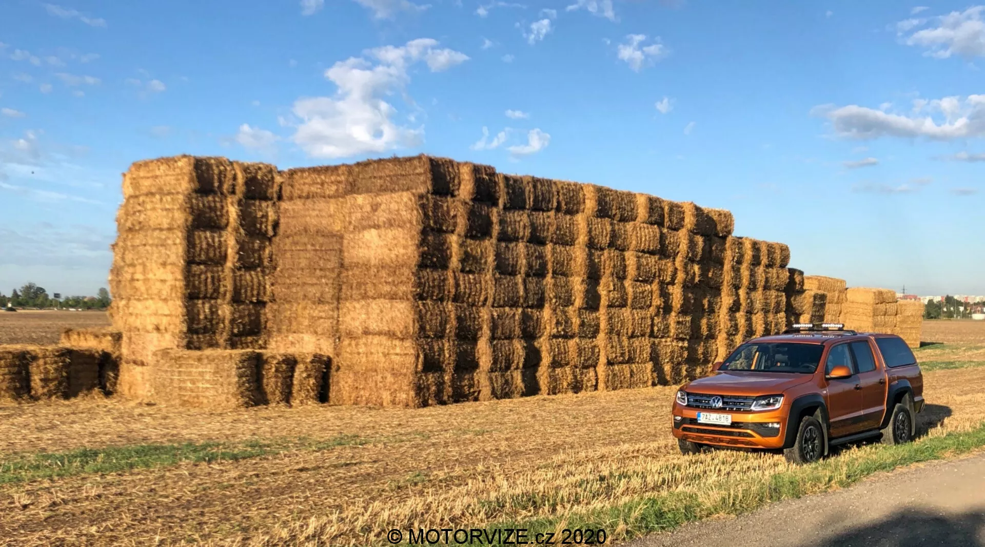 Um Volkswagen Amarok 2016 estacionado em frente a grandes fardos de feno em ambiente rural. A perspectiva de três quartos frontal captura a grade, os faróis, o para-choque dianteiro, o capô, as rodas, os espelhos laterais e as portas do carro. A proeminente grade de ripas horizontais e os faróis angulares acentuados melhoram a aparência contemporânea do veículo. As janelas escurecidas, os trilhos do teto e os painéis de proteção ao redor das portas sugerem versatilidade e capacidade robusta. O veículo está situado contra um fundo de fardos de feno, acrescentando contexto para seu uso em estradas rurais ou na fazenda. A luz da manhã ou do final da tarde projeta sombras quentes e realça a cor e os detalhes do design do carro. Com marca d'água 'MOTORVIZE.cz 2020'.