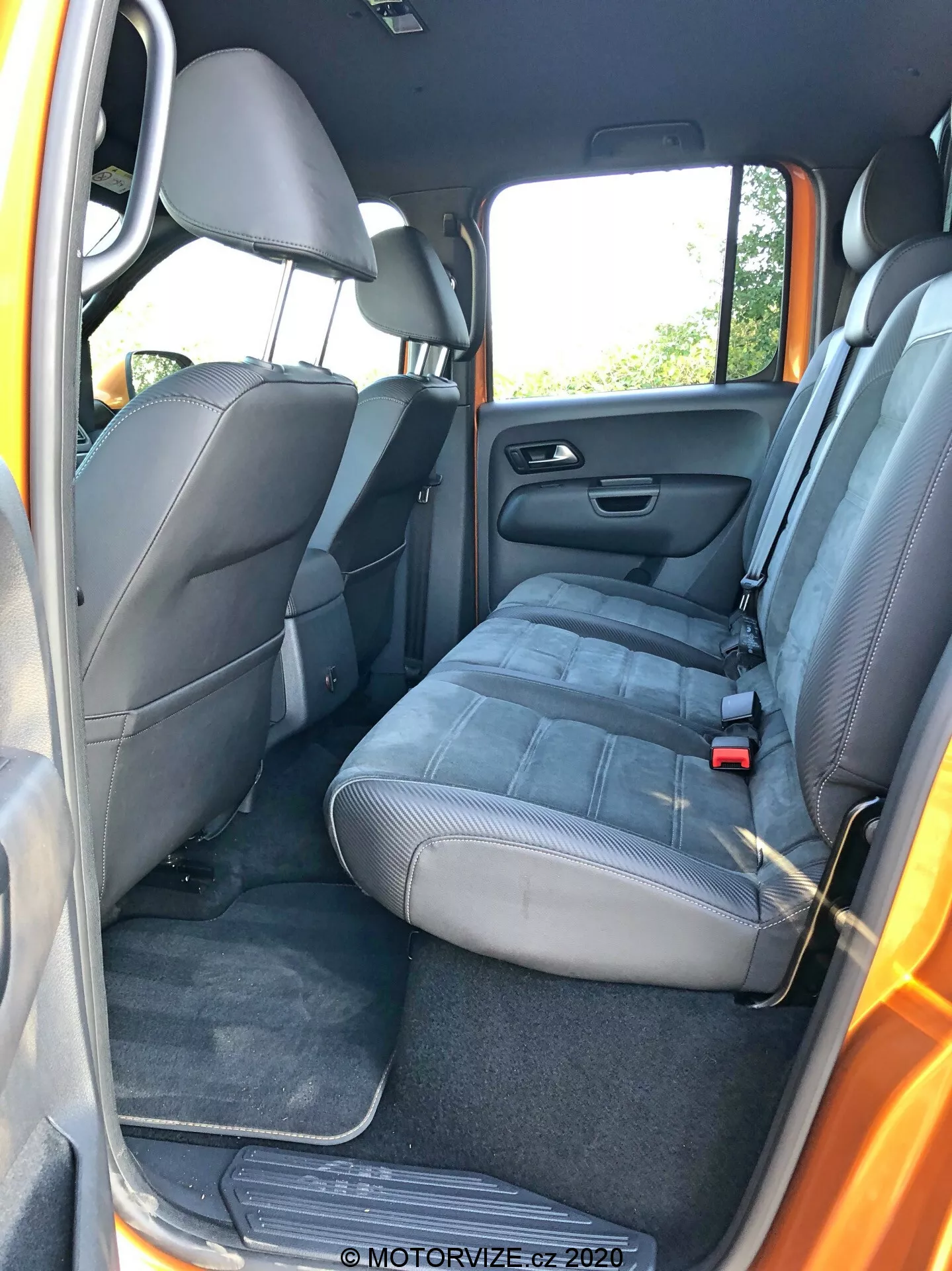 Vista interior do VW-Nutzfahrzeuge Amarok 2016 a partir de um ângulo da porta traseira do passageiro; mostra um banco traseiro em estilo banco adequado para três passageiros com estofamento em tecido, apoios de cabeça, cintos de segurança (cinto abdominal no centro, cintos de três pontos nos lados), porta aberta revelando painel da porta com alto-falante, armazenamento e controle do vidro elétrico; piso acarpetado e bancos traseiros rebatíveis; bolso de armazenamento nas costas do banco do passageiro dianteiro; características padrão do carro sem elementos de alta tecnologia notáveis; luz do dia natural.