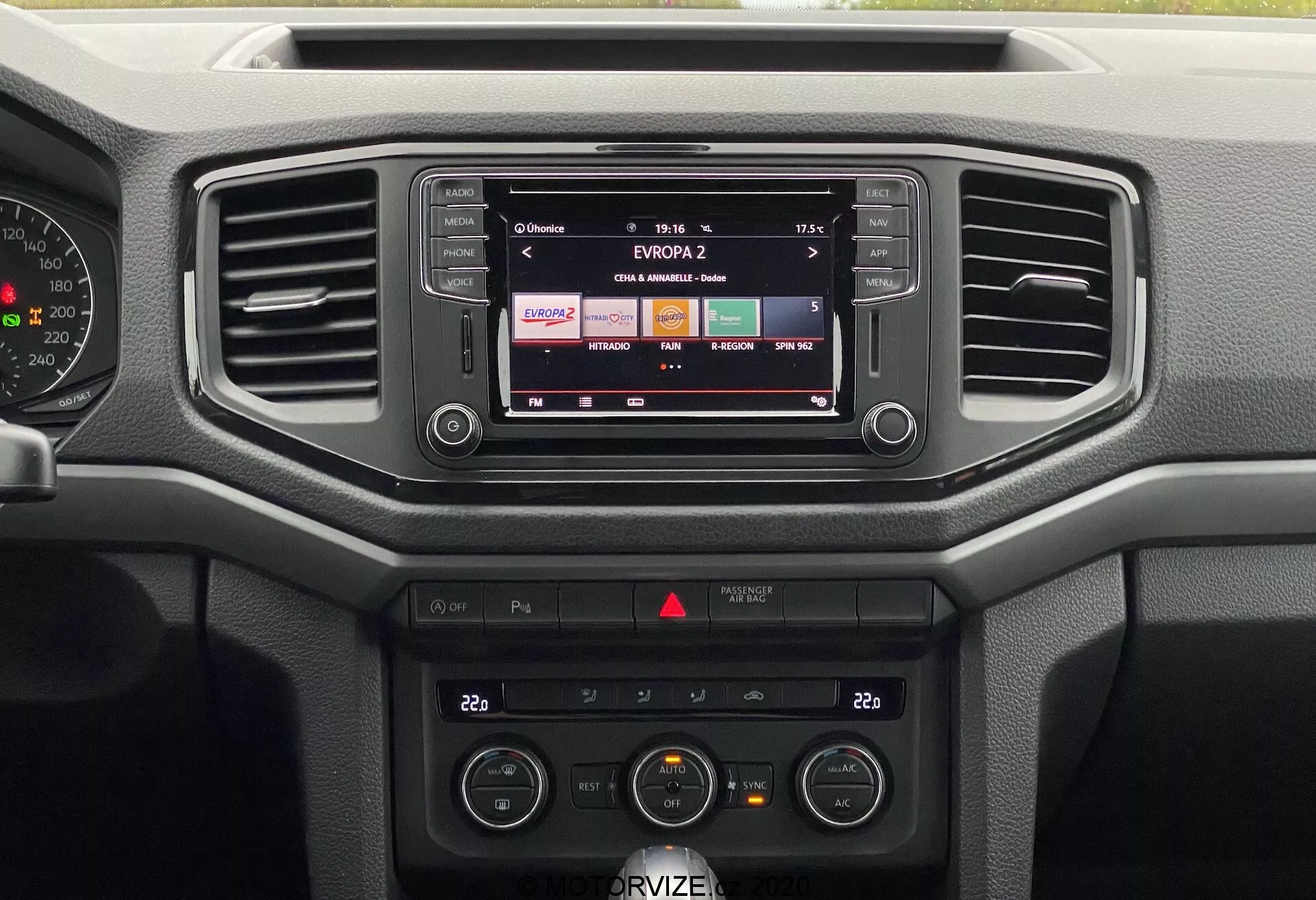 A imagem apresenta o painel central do VW Nutzfahrzeuge Amarok 2016. De uma perspectiva frontal, você pode ver o sistema de infotainment exibindo uma estação de rádio, dois ventiladores horizontais e vários controles. O sistema de áudio e o controle de clima, incluindo os ajustes de temperatura e ventilador, são claramente visíveis. O painel prioriza a funcionalidade com botões e mostradores fáceis de usar, utilizando principalmente materiais plásticos rígidos, adequados para uso diário em vez de luxo.