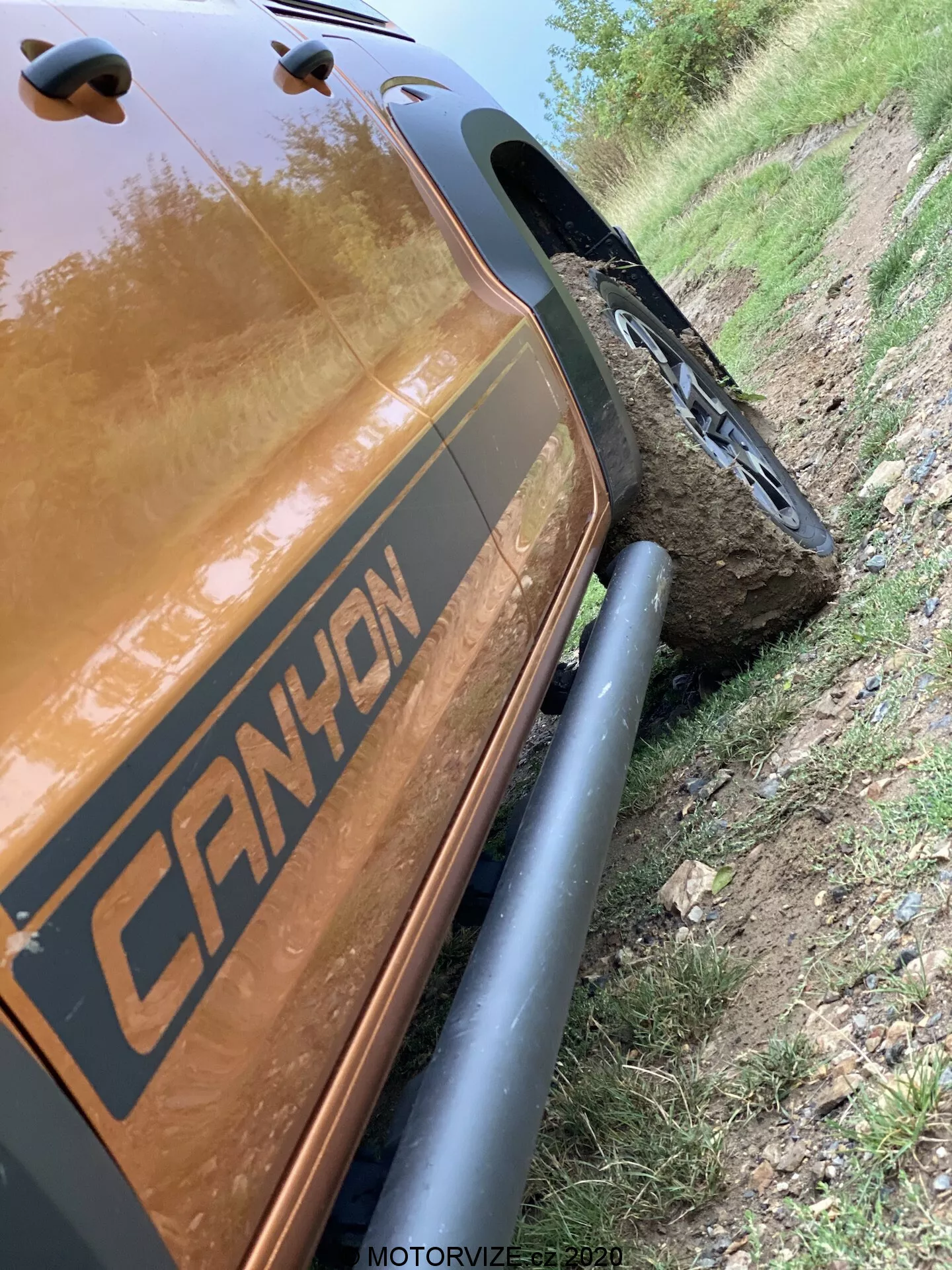 Esta imagem de ângulo inclinado a 45 graus mostra a seção traseira do lado do passageiro de um VW-Nutzfahrzeuge Amarok 2016. O foco está nos detalhes próximos da cava da roda traseira até a seção de carga traseira, destacando os robustos arcos protetores de roda pretos e os robustos pneus off-road. O veículo apresenta um gráfico com a inscrição 'CANYON' e está localizado ao lado de um caminho de terra com uma roda montada em uma grande pedra, demonstrando sua capacidade off-road. A cor metálica refletiva marrom ou cobre do corpo do carro é evidente, capturando reflexos de luz. A marca de tempo 'MOTORVIZE.cz 2020' sugere que foi capturada para uma publicação de veículos motorizados.