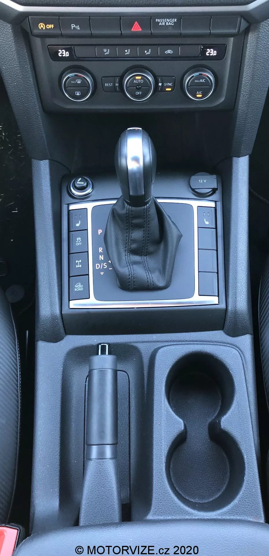 Esta imagem mostra o console central de um vw-nutzfahrzeuge amarok 2016, vista de uma perspectiva aérea aproximadamente do ponto de vista do motorista ou do passageiro da frente. A fotografia captura o interior do carro especificamente para a alavanca de câmbio, controles do console central e compartimentos de armazenamento. A imagem inclui mostradores e botões do sistema de controle climático, uma alavanca de câmbio revestida em couro em um painel prateado indicando transmissão automática, um controle eletrônico de freio de estacionamento, um compartimento de armazenamento, dois porta-copos e armazenamento adicional em direção aos assentos traseiros. Os materiais visíveis incluem plástico, acentos metálicos e couro, refletindo uma combinação de funcionalidade e estética.