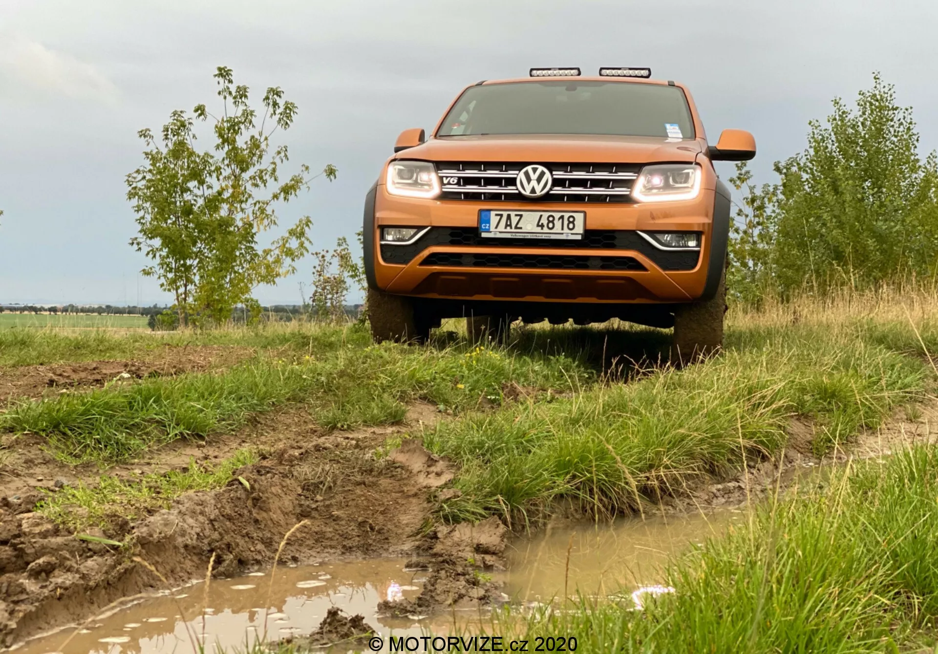 Uma vista frontal, em ângulo ligeiramente baixo, de uma Volkswagen Amarok 2016 laranja, uma caminhonete de médio porte, conhecida por suas capacidades off-road. A imagem captura a frente do veículo, incluindo o capô, a grade, o para-choque, parte dos para-lamas, os faróis e o logotipo da Volkswagen exposto no centro da grade. Acima da grade, há detalhes cromados ou luzes diurnas de LED. O terreno lamacento e a grama ao redor do carro enfatizam sua natureza off-road, com luzes adicionais no teto, uma placa de deslizamento frontal e um pano de fundo rural com céu nublado completam o ambiente aventureiro. A imagem é capturada a uma distância de aproximadamente dois a três metros, proporcionando uma visão detalhada das características frontais do veículo e sua interação com o ambiente.
