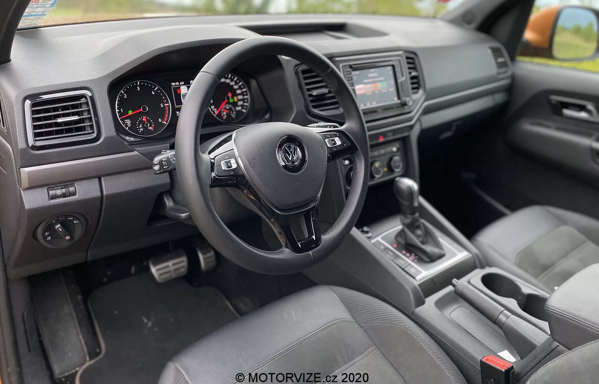 Vista interior do VW Nutzfahrzeuge Amarok 2016 do lado do passageiro, mostrando layout de volante à direita com volante Volkswagen de três raios com botões de controle, painel de instrumentos com velocímetro e tacômetro analógicos, console central com sistema de áudio e tela de infoentretenimento, alavanca de câmbio na posição 'P', assentos estofados em tecido, painel com saídas de ar e plásticos texturizados, e painel da porta do motorista com controle de vidros elétricos e ajustes de espelhos.