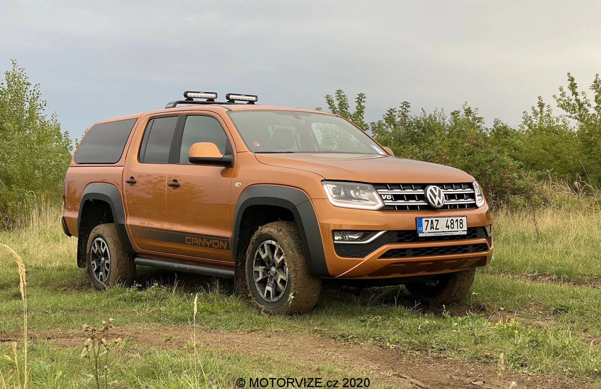 A imagem mostra uma pickup Volkswagen Amarok 2016 laranja estacionada em um terreno gramado irregular com um céu nublado ao fundo. A perspectiva lateral-frontal se concentra no lado do motorista, mostrando o perfil do veículo e parte da frente. As principais características visíveis incluem um bagageiro de teto, rodas cobertas de lama, indicando atividade off-road recente, degraus laterais, configuração de cabine dupla, design moderno de faróis e um para-choque dianteiro com faróis de neblina e placa de proteção inferior. O veículo exala uma estética utilitária robusta para off-road, com elementos tecnológicos modernos.