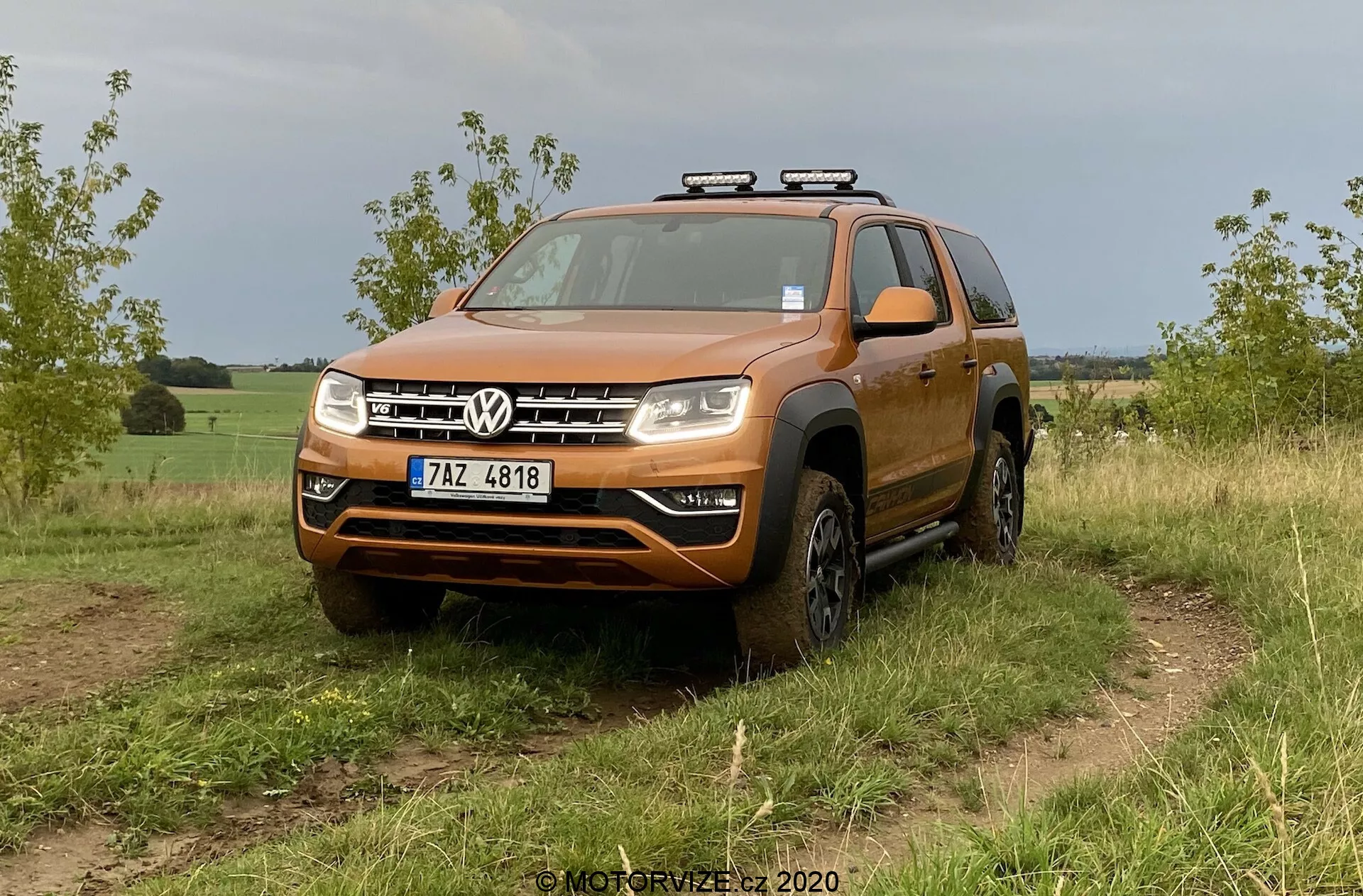 Um SUV Volkswagen laranja, especificamente o VW Amarok 2016, é exibido em um terreno off-road coberto de grama. A foto, tirada de um ângulo frontal baixo a médio, enfatiza seu design robusto, adequado para atividades off-road. O veículo possui uma grade frontal ousada com duas barras cromadas emoldurando o logotipo central da Volkswagen, um para-choque de tamanho considerável com uma possível placa protetora, faróis compostos claros com potenciais lâmpadas LED ou halógenas e arcos de roda alargados que abrigam pneus off-road. As luzes auxiliares estão montadas no teto, sugerindo funcionalidade adicional para condução noturna. Detalhes visíveis incluem o para-brisa, trilhos de teto, espelhos laterais com indicadores de direção integrados e uma placa de licença frontal. O veículo parece limpo com um pouco de sujeira nos lados inferiores e nas rodas, o que é compatível com o uso off-road. O fundo sugere um ambiente rural ou semi-rural.