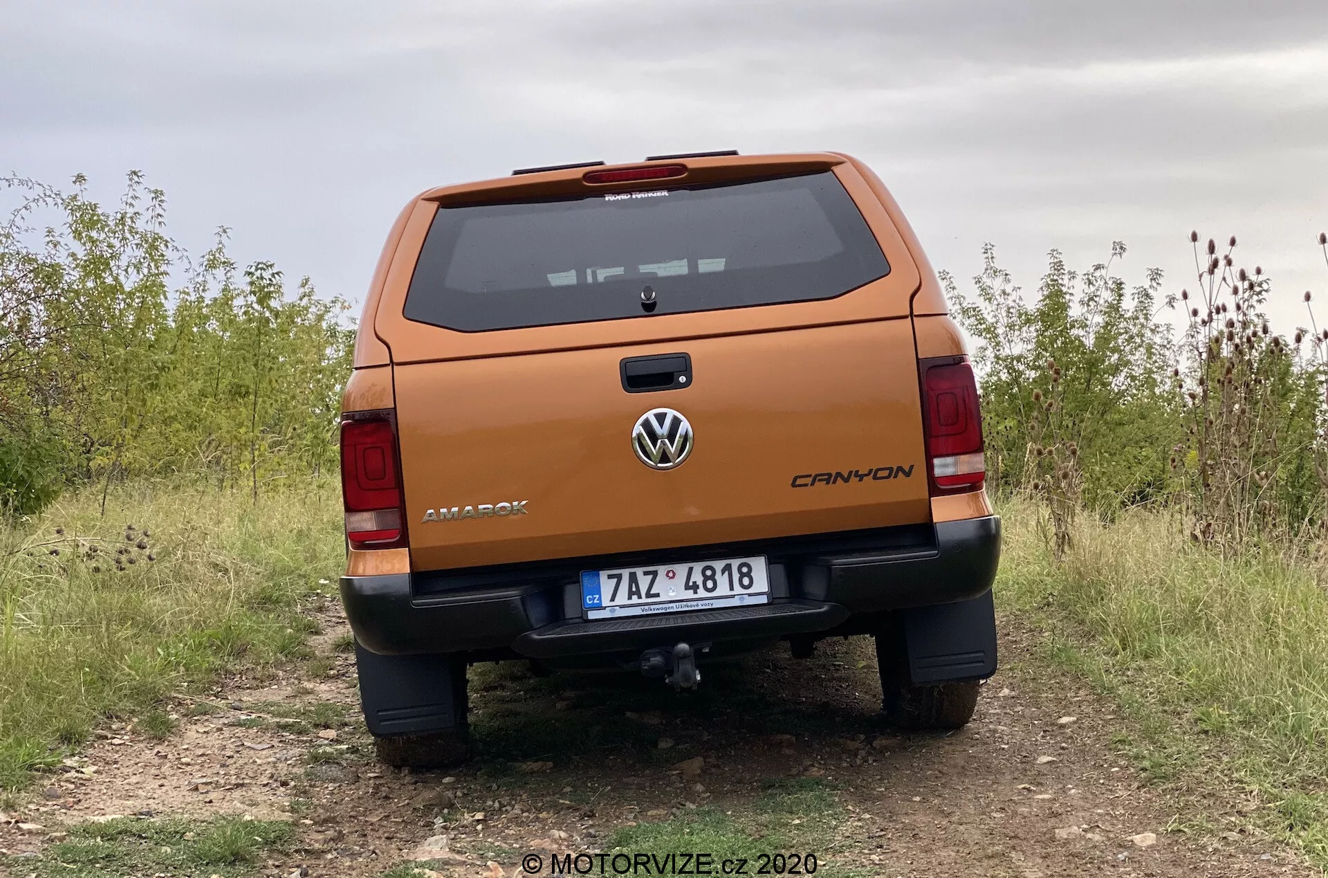 Vista traseira profissional de uma picape Volkswagen Amarok 2016 a partir de um ângulo baixo, enfatizando a robusta tampa traseira com a marca 'AMAROK', logotipo da Volkswagen centralizado, indicação do nível de acabamento 'CANYON', placa de licença, luzes traseiras detalhadas, para-choque com sensores de estacionamento e degrau, barra de reboque, estacionada em um caminho não pavimentado indicando capacidade off-road.