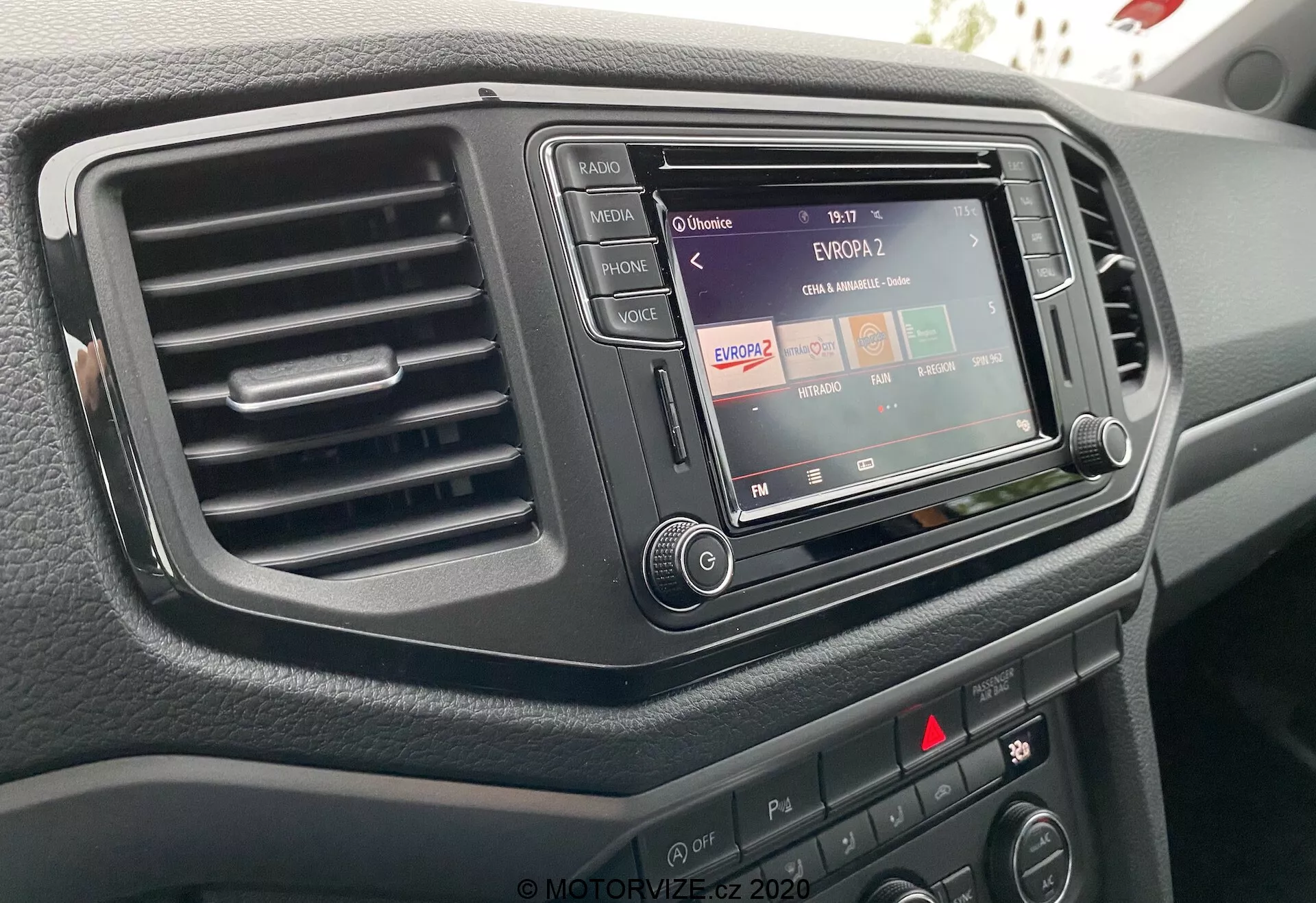 Close-up do console central de um VW Nutzfahrzeuge Amarok 2016 a partir da perspectiva do motorista, destacando o sistema de infoentretenimento com funcionalidade de rádio, uma única saída de ar à esquerda e vários botões e manípulos de controle. O sistema de infoentretenimento está embutido em um acabamento escuro e elegante, e a superfície do painel tem um acabamento texturizado.
