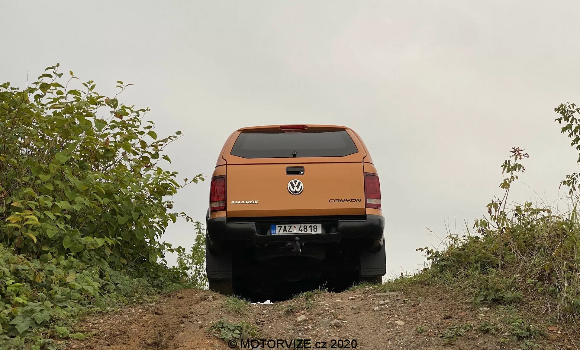Vista traseira de um crossover ou SUV Volkswagen laranja de um ângulo ligeiramente baixo, capturada no topo de uma pequena colina. Os elementos de design traseiro do veículo, como a porta traseira, a janela traseira, as luzes traseiras, as emblemas da marca e do modelo que leem 'Volkswagen' e 'CROSSOVER,' e o para-choque traseiro são claramente visíveis. A placa de licença é visível, mas detalhes específicos não são fornecidos. A porta traseira apresenta um vinco proeminente e linhas de design, com uma possível alça ou recesso abaixo da placa de registro. As luzes traseiras envolvem para os lados e incluem indicadores, luzes de ré e luzes de freio. A parte inferior preta do para-choque traseiro sugere um acabamento protetor para uso off-road. O veículo é emoldurado por vegetação, indicando um ambiente rural ou off-road. O chão é de solo natural, consistente com as capacidades off-road do veículo. No canto inferior direito, há uma marca d'água que lê 'MOTORVIZE.cz ©2020'.