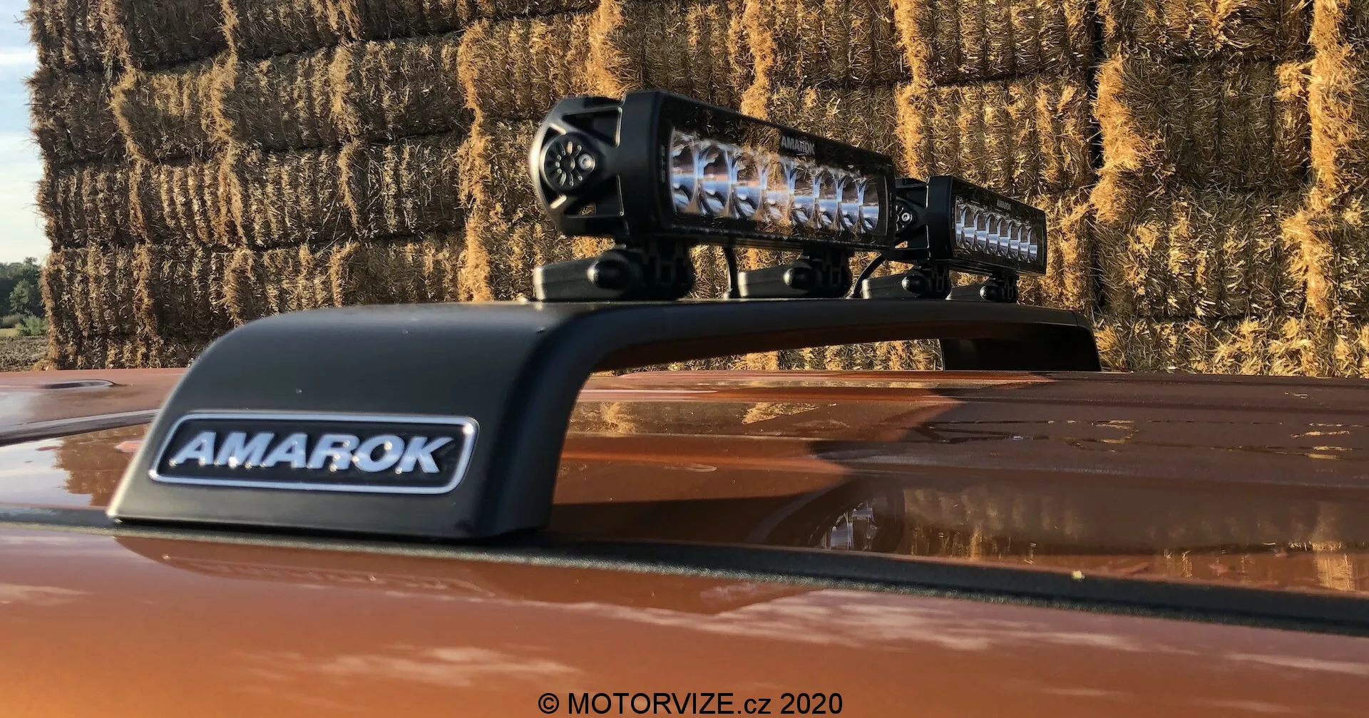 Close-up de uma picape Volkswagen Amarok 2016 mostrando uma barra de luz LED aftermarket montada no teto. O invólucro preto da barra de luz com elementos LED individuais é claramente visível, com a marca 'AMAROK' em uma cobertura ventilada. A foto, tirada ao ar livre, mostra as lentes reflexivas da barra de luz e fardos de feno ao fundo, indicando um ambiente rural.