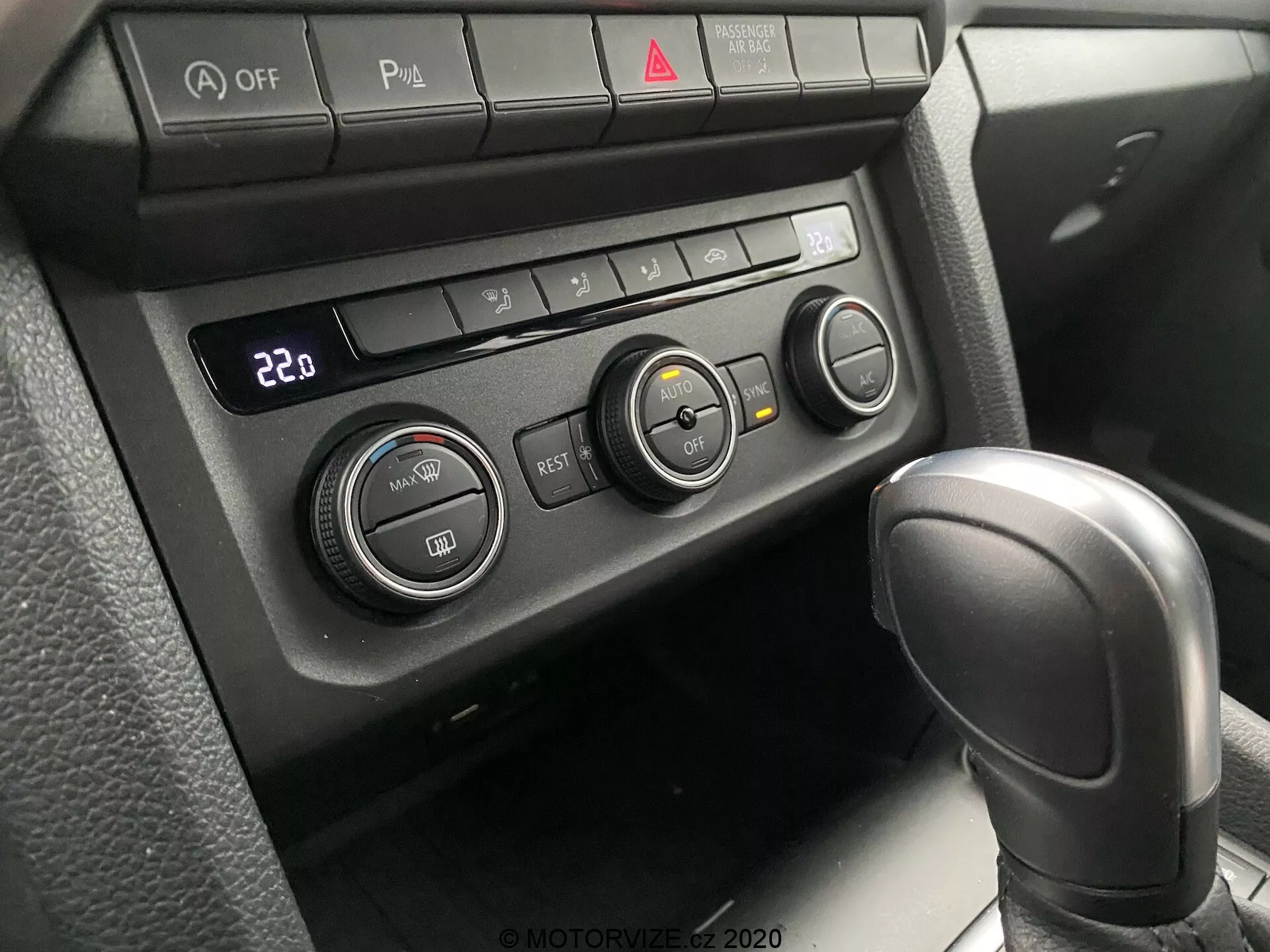 Close-up do sistema de controle climático e parte da alavanca de câmbio do VW Nutzfahrzeuge Amarok 2016. Perspectiva do passageiro ou motorista olhando para baixo em direção ao console central. Apresenta uma alavanca de câmbio automática texturizada em couro em 'Park' e um painel de controle climático ajustado para 22°C com três botões rotativos e vários botões funcionais. Botões incluem desativação do Auto Stop/Start, comutador de sensores de estacionamento, volante aquecido e aquecimento de assento, juntamente com botão de luz de emergência vermelho central. A iluminação suave e difusa sugere um interior bem sombreado.