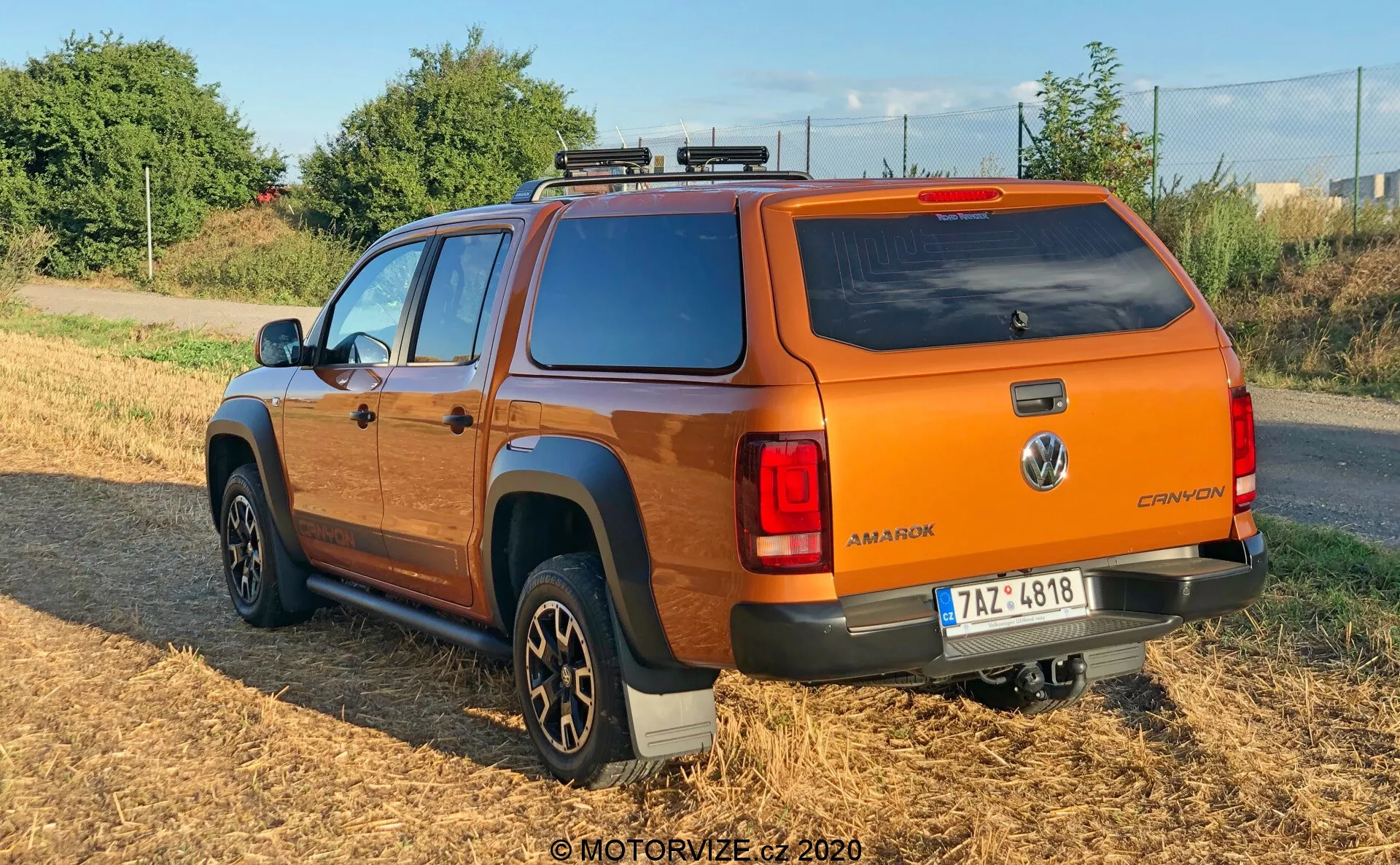 Vista traseira de três quartos de uma picape Volkswagen Amarok 2016 laranja com capota rígida, sistema de bagageiro de teto e rodas de liga leve de múltiplos raios. O veículo está estacionado em uma área de grama seca com iluminação natural suave, exibindo o emblema da Volkswagen, nome do modelo 'AMAROK' e designação 'CANYON', e luzes traseiras modernas. As estribos laterais de cor escura e a configuração de cabine dupla ressaltam sua funcionalidade utilitária e de passageiros.