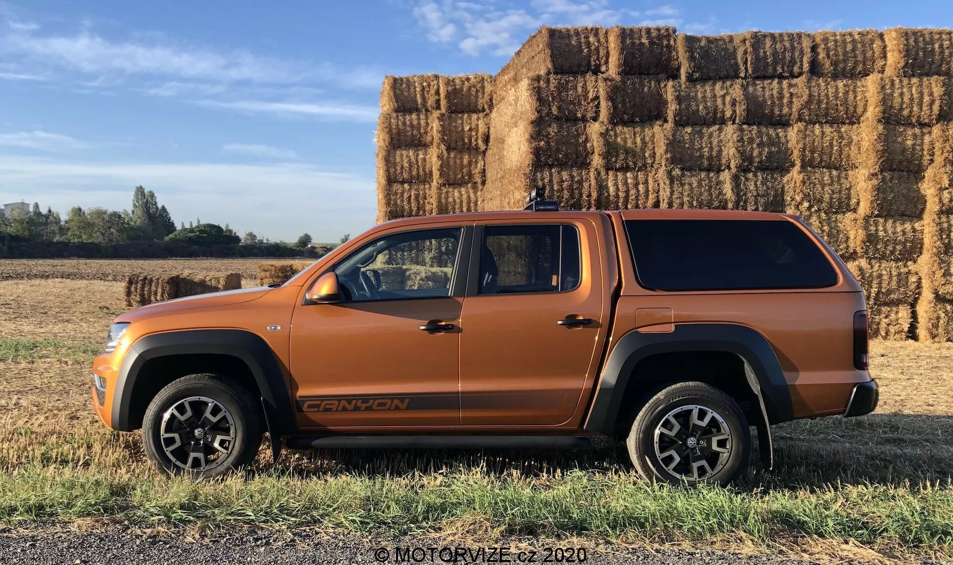 Vista de perfil lateral de um veículo tipo SUV metálico laranja, provavelmente um SUV médio ou pickup com cabine estendida e cobertura da caçamba, estacionado em um terreno plano e gramado com grandes fardos de feno retangulares empilhados ao fundo sob um céu claro e ensolarado. O veículo apresenta linhas de carroceria arrojadas, grandes arcos de roda, acabamento inferior preto, robustas rodas de liga leve, emblema visível na parte inferior da porta dianteira e antena no teto. A fotografia destaca as possíveis capacidades off-road e o design contínuo da cobertura da caçamba.