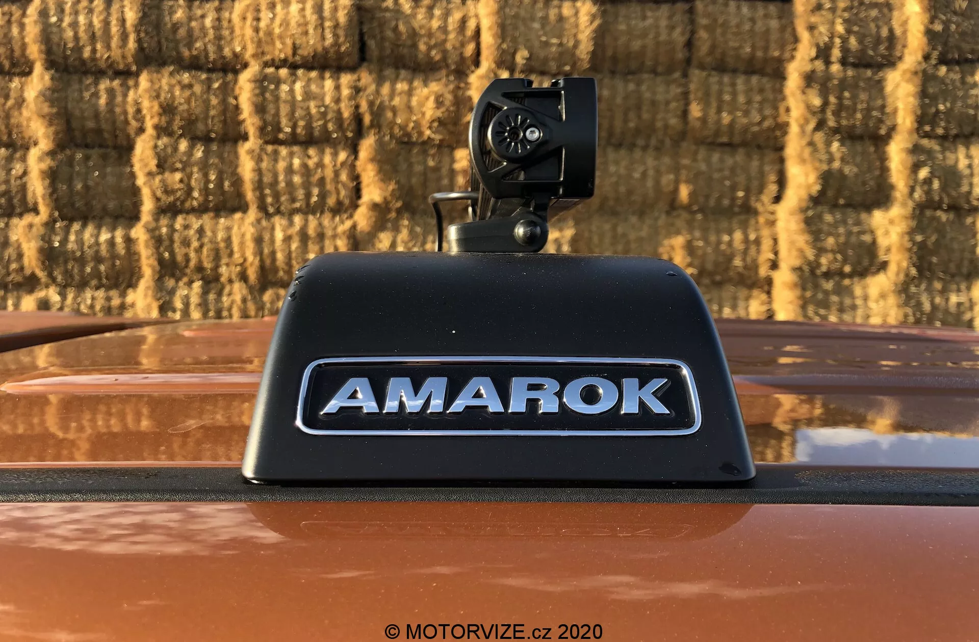 Foto em close-up de um caminhão pickup Volkswagen Amarok 2016 com uma câmera de ré instalada no alojamento da luz de freio montada alta com 'AMAROK' em letras prateadas em relevo sobre uma carroceria metálica de cor bronze ao ar livre contra um fundo com padrão de tijolos.