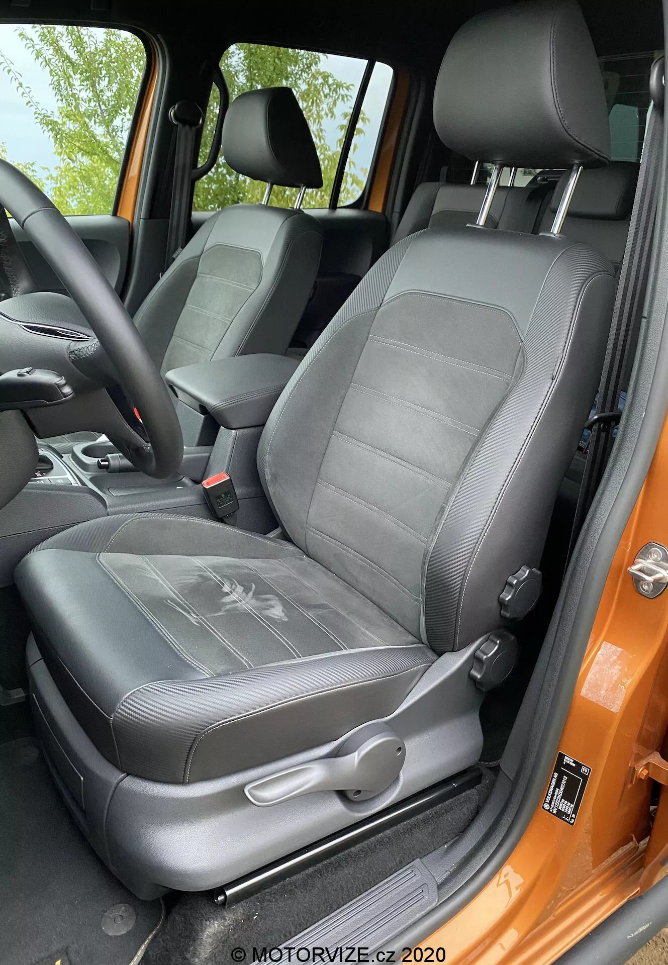 Vista aproximada do banco do motorista do VW-nutzfahrzeuge Amarok 2016 a partir da perspectiva de alguém parado fora da porta aberta. O banco revestido em material semelhante a couro preto apresenta um design texturizado com costuras em relevo e apoio de cabeça integrado ajustável. Visíveis controles manuais de ajuste e o mecanismo do cinto de segurança de três pontos. O suporte lateral indica suporte lateral, e a moldura da porta pintada em cor laranja vibrante contrasta com o interior preto. O banco parece bem conservado e iluminado por luz natural.