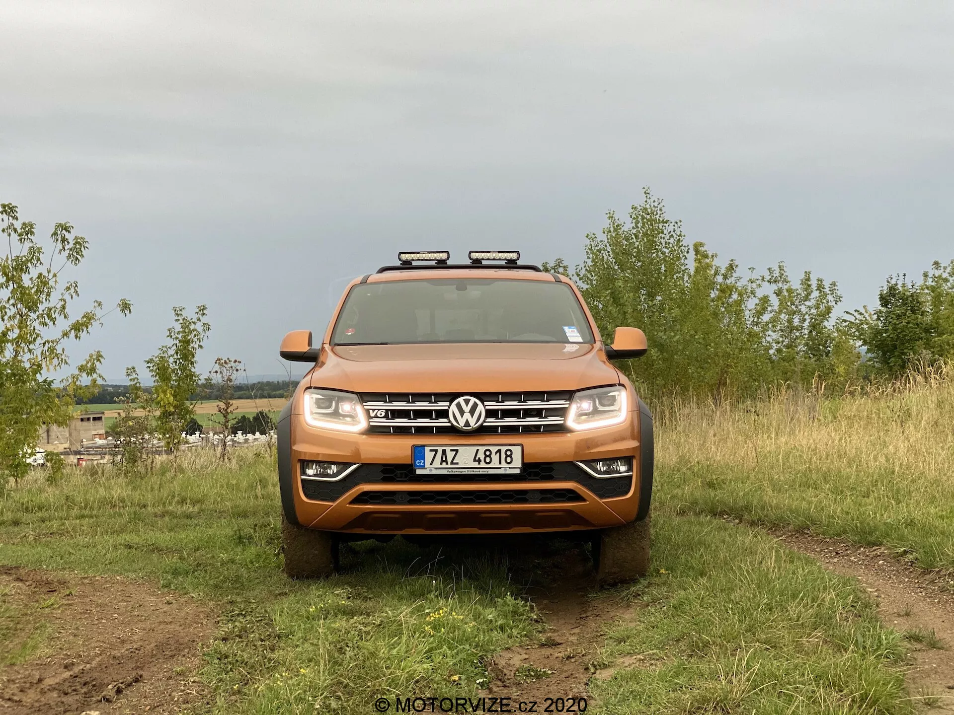 Vista frontal de um SUV Volkswagen Amarok capturada em um ambiente externo. O veículo laranja apresenta emblema Volkswagen proeminente na grade, lâminas horizontais, faróis padrão e luzes auxiliares adicionais montadas no teto. A imagem destaca as capacidades off-road com pneus robustos parcialmente visíveis e sujeira nas superfícies do veículo. Está posicionado em um terreno natural com fundo desfocado, iluminado difusamente por um céu nublado.