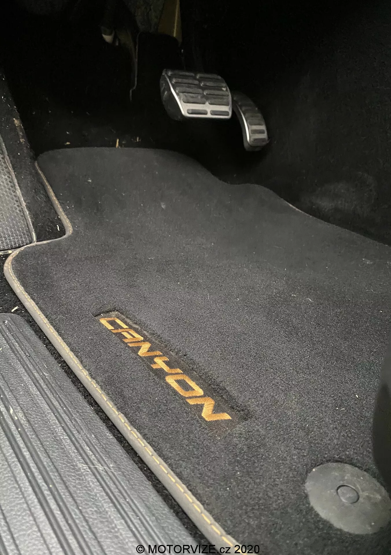 Close-up da área do piso do lado do motorista no VW-Nutzfahrzeuge Amarok 2016, mostrando um tapete escuro com bordado 'ENYON'. São visíveis os pedais de acelerador e freio metálicos, com o pedal de freio em posição superior, sugerindo que está acionado. O tapete é contornado para se ajustar ao espaço, com um gancho de retenção à direita. O tapete contrasta com o ambiente mais escuro, indicando um interior de temática escura. A cena é fracamente iluminada, provavelmente tirada à noite ou em condições de pouca luz.