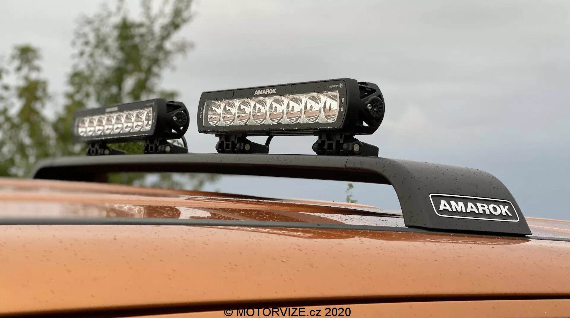 Close-up focado para cima do teto de um Volkswagen Amarok 2016 mostrando equipamento de iluminação montado no teto com duas barras de luz LED de alta intensidade em um suporte com a marca 'AMAROK', acabamento de pintura metálica laranja; ideal para características de veículos off-road ou acessórios para veículos.