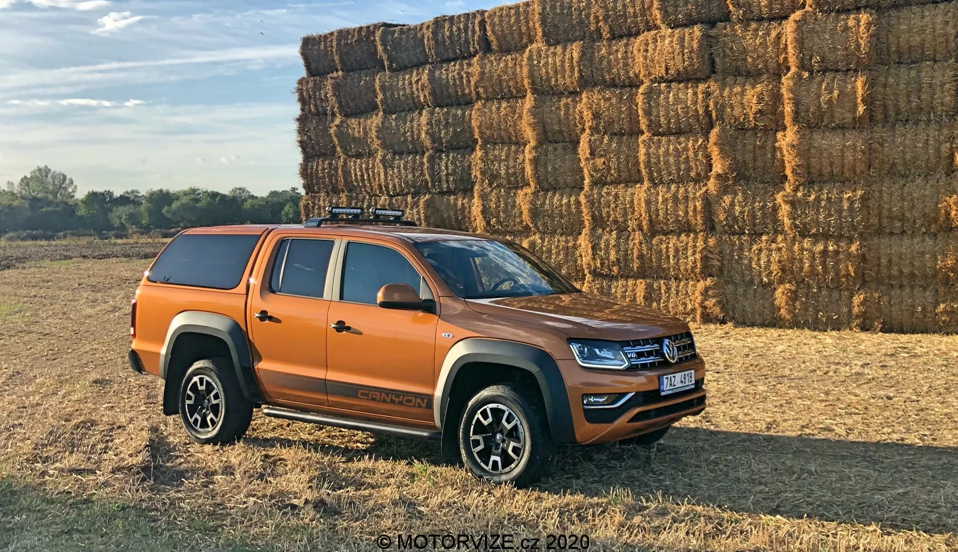 Vista lateral de uma pick-up Volkswagen Amarok laranja estacionada ao ar livre em uma superfície irregular, possivelmente de terra ou grama. O veículo está posicionado em um ângulo de 30 graus em relação à câmera, mostrando o perfil lateral e uma visão parcial da grade frontal e dos faróis. A moderna pick-up de tamanho médio apresenta linhas de design frescas e iluminação LED, indicando um modelo recente. Visíveis são quatro portas indicando uma configuração de cabine dupla, rodas de liga leve multirraio, pneus off-road, uma cobertura rígida na caçamba do caminhão e um sistema de rack de teto. Os espelhos laterais provavelmente incluem indicadores. Fotografado à luz do dia quente com sombras longas, uma grande pilha de fardos de feno ao fundo sugere um ambiente rural.
