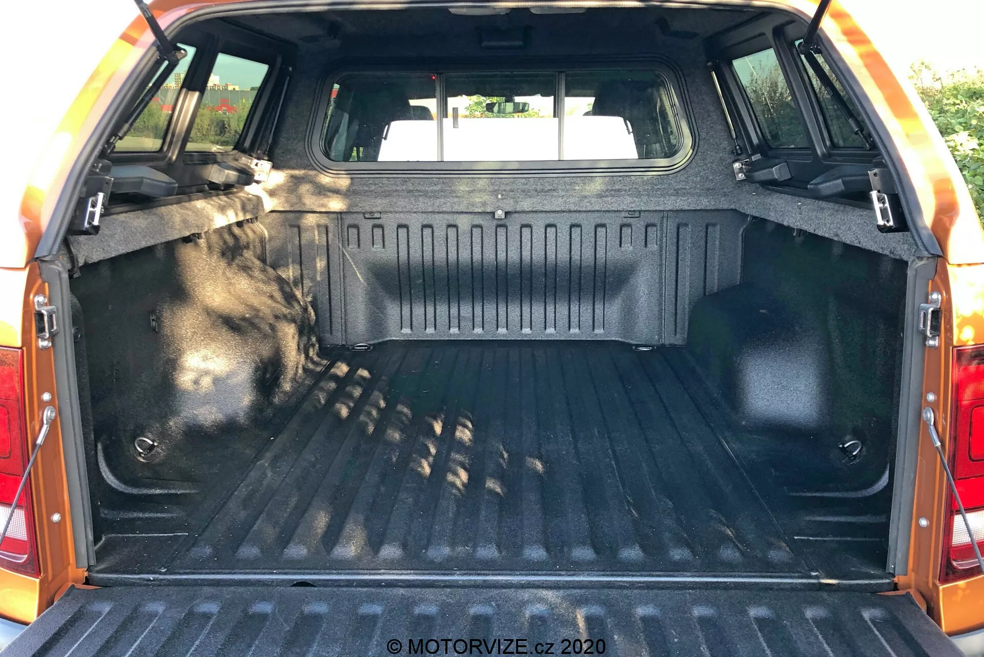 Interior do espaço de carga do VW Nutzfahrzeuge Amarok 2016, visto da parte traseira com a tampa do porta-malas aberta, mostrando a área do porta-malas. A área de carga possui um revestimento robusto e texturizado, paredes laterais acabadas com material durável, pontos de amarração para segurar itens, iluminação interna para visibilidade, bancos traseiros visíveis na parte frontal do espaço e borda protetora na borda da tampa do porta-malas.