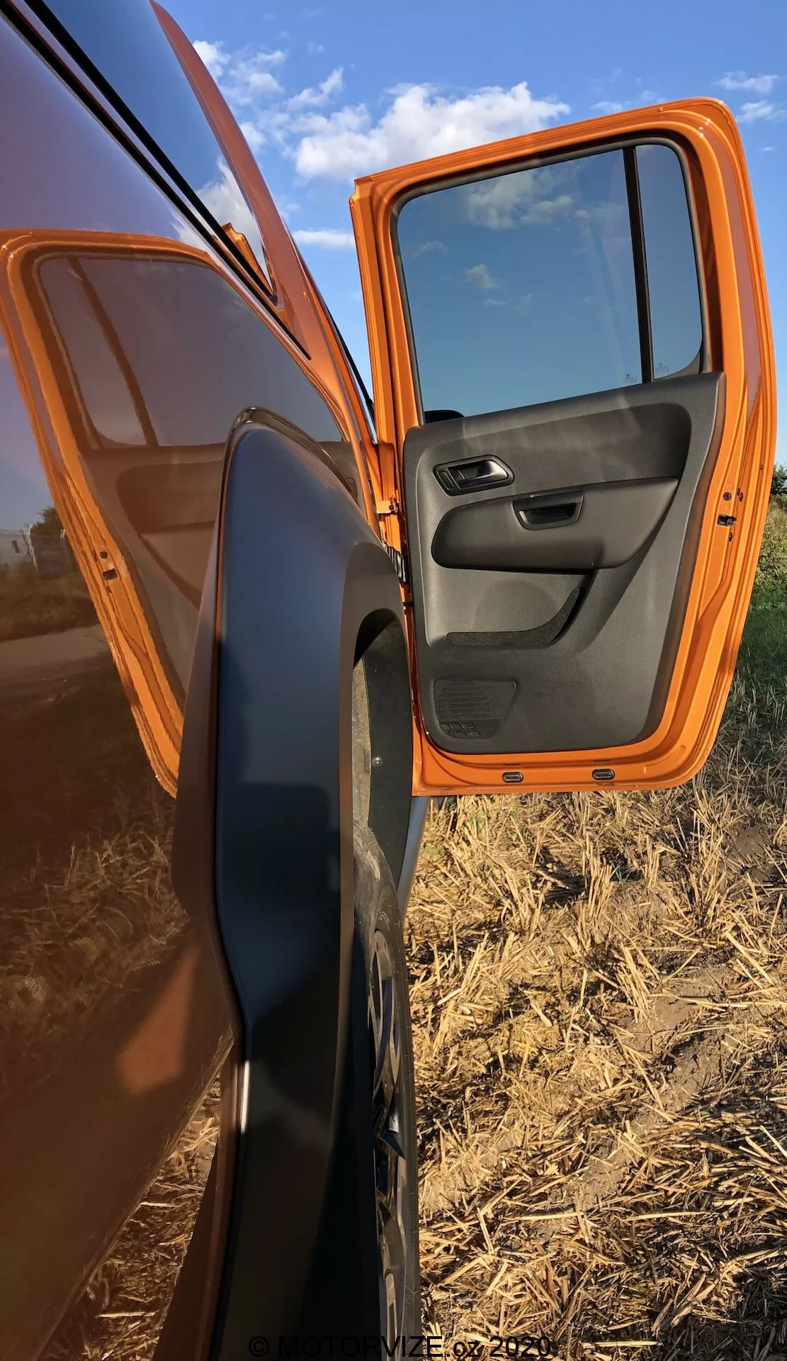 Vista aproximada de uma porta da frente do passageiro parcialmente aberta de um VW Nutzfahrzeuge Amarok 2016 laranja, mostrando a pintura externa laranja brilhante e o painel interno preto contrastante com controles integrados. O carro está estacionado diagonalmente, apresentando um perfil lateral com a porta aberta em um ângulo de 70 graus, destacando o espelho lateral com caixa preta. Os reflexos na porta mostram um céu claro e campos agrícolas planos, acentuando o ambiente rural.