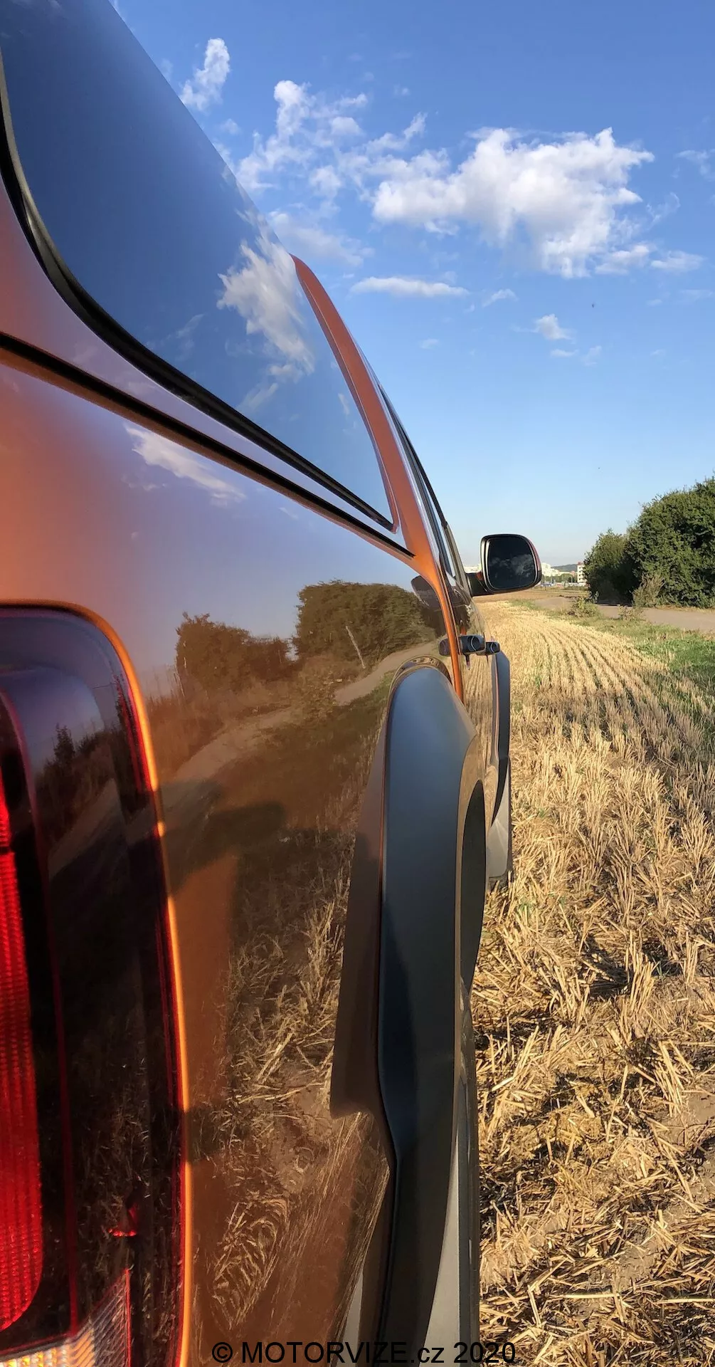 Vista lateral aproximada do painel traseiro direito de um VW Nutzfahrzeuge Amarok 2016, destacando a carroceria metálica brilhante e refletiva e a lanterna traseira integrada com lente clara. A foto, tirada de um ângulo baixo, mostra a linha de cintura, o acabamento de plástico preto e o campo colhido rural ao fundo com um céu azul claro.