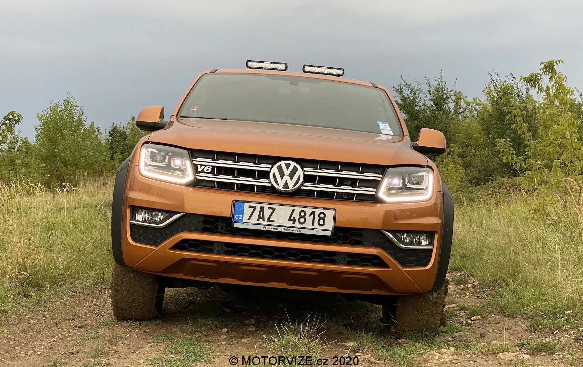 Vista frontal de um pick-up Volkswagen Amarok laranja metálico, mostrando sua postura robusta e capacidades off-road com uma grade proeminente com o emblema VW e insígnia V6. A imagem, tirada de um ângulo baixo, captura toda a largura da frente do veículo, incluindo os faróis e luzes adicionais off-road, com um fundo natural de um campo e árvores destacando a robustez do pick-up. As características visíveis incluem para-choques dianteiro robusto, placa de proteção, sistema de iluminação avançado ativado e placa de matrícula da República Tcheca.