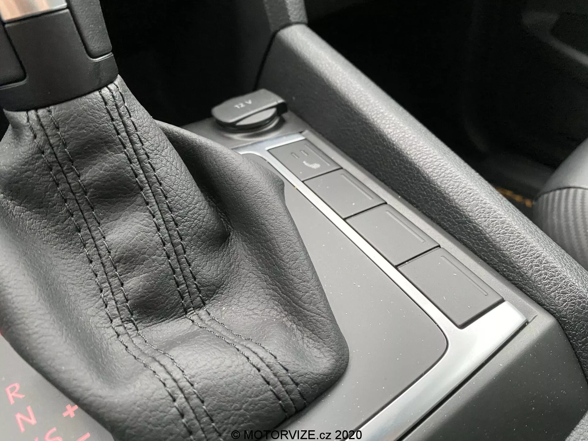 Imagem em close-up do console central de um VW Nutzfahrzeuge Amarok 2016, focando na área da alavanca de câmbio. A alavanca de câmbio, coberta em couro preto com costuras visíveis, é o ponto principal. As letras 'PRNDS' atrás da alavanca indicam uma transmissão automática. À direita há quatro botões quadrados nivelados com a superfície do console, possivelmente controlando várias funções do carro. A área atrás dos botões pode ser um pequeno espaço de armazenamento. Os materiais e qualidade de construção sugerem um alto nível de artesanato. A luz ambiente do canto superior esquerdo destaca os detalhes.