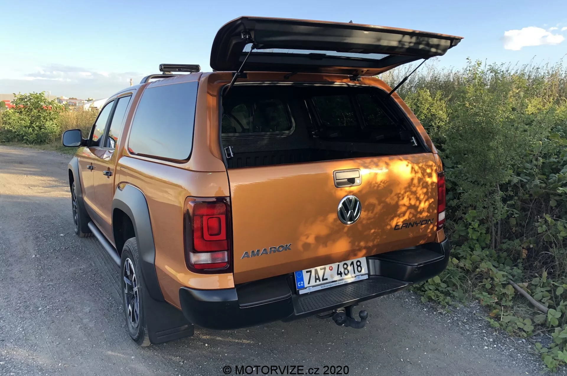 Vista traseira de três quartos de uma caminhonete Volkswagen Amarok 2016 em laranja metálico, destacando a tampa da caçamba aberta, o engate de reboque, as portas da cabine dupla da segunda fila e parte da roda traseira direita e do para-choque, ambientada em um ambiente rural ao entardecer. Foto por MOTORVIZE.cz, 2020.