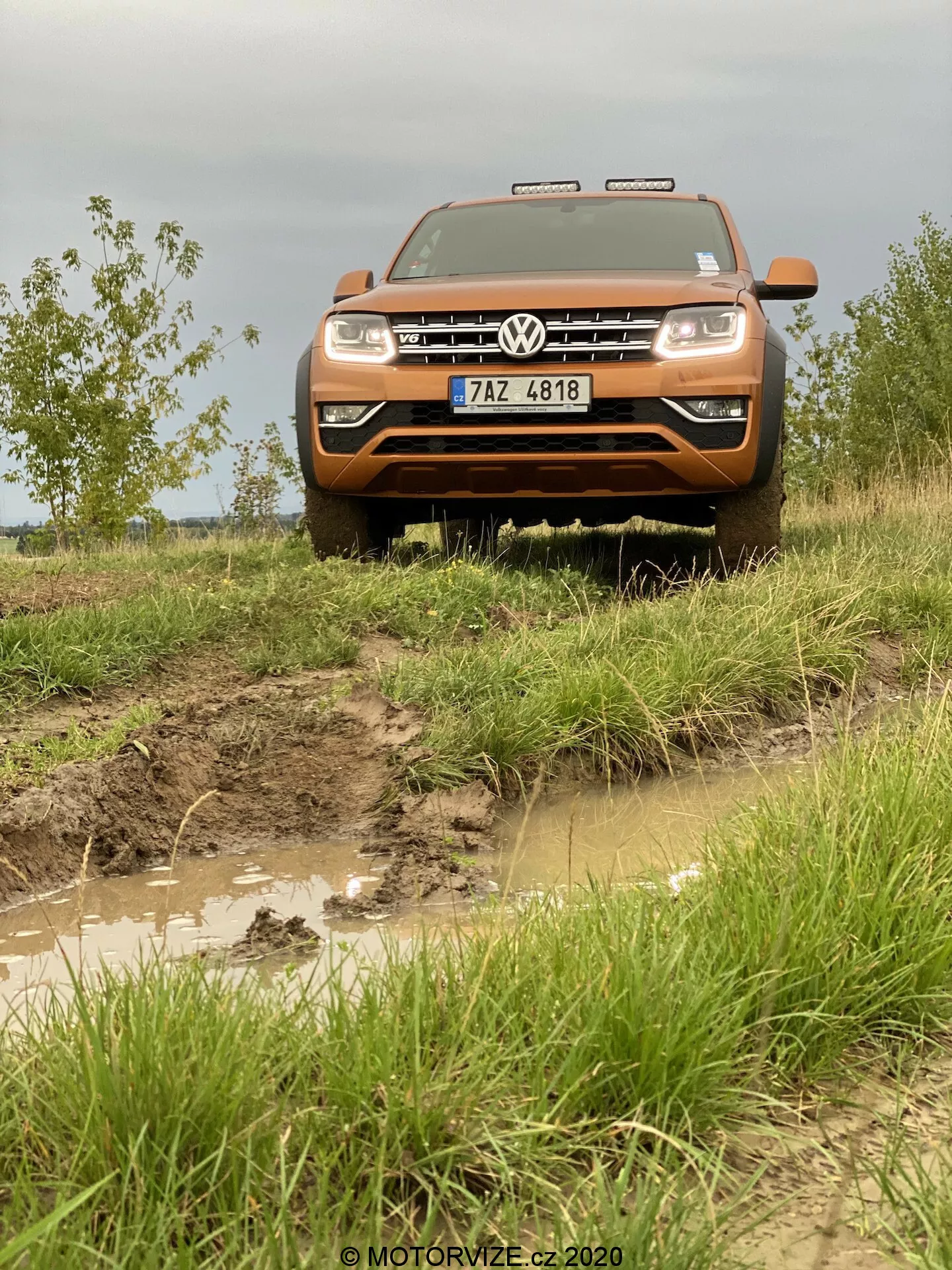 Vista frontal de um veículo off-road Volkswagen Amarok 2016 laranja, destacando a grade frontal, pára-choques, capô e conjunto de faróis. Fotografada de um ângulo baixo para enfatizar sua postura elevada, a imagem revela uma trilha acidentada e lamacenta com poças de água e exterior salpicado de lama, acentuando suas capacidades off-road. O veículo apresenta a grade Volkswagen proeminente com emblema, faróis retangulares e iluminação auxiliar acima da grade. A placa de licença é visível no robusto pára-choques dianteiro, enquanto o capô plano e alongado destaca o tamanho e potência do Amarok.