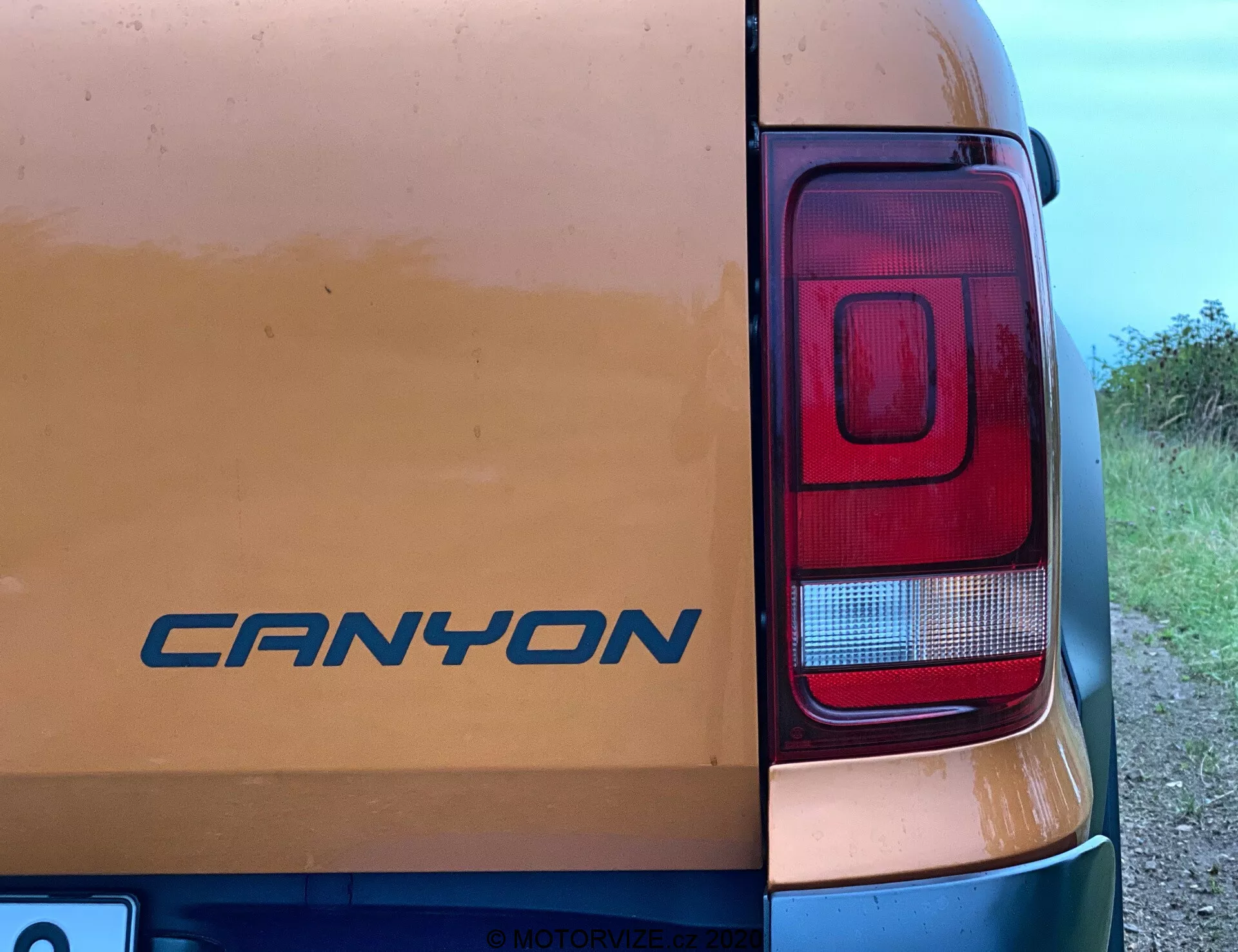 Vista aproximada do canto traseiro direito de um veículo VW Nutzfahrzeuge Amarok 2016. O emblema 'CANYON' é visível na porta traseira em meio a sinais de sujeira leve, sugerindo uso em estradas de terra. A fotografia destaca principalmente o conjunto de luzes traseiras retangulares e modernas com seções para freio, ré e possivelmente sinalizadores de direção. Tirada de um ângulo lateral aproximado de 45 graus, a imagem captura parcialmente a carroceria do veículo com um fundo rural ou semi-rural.