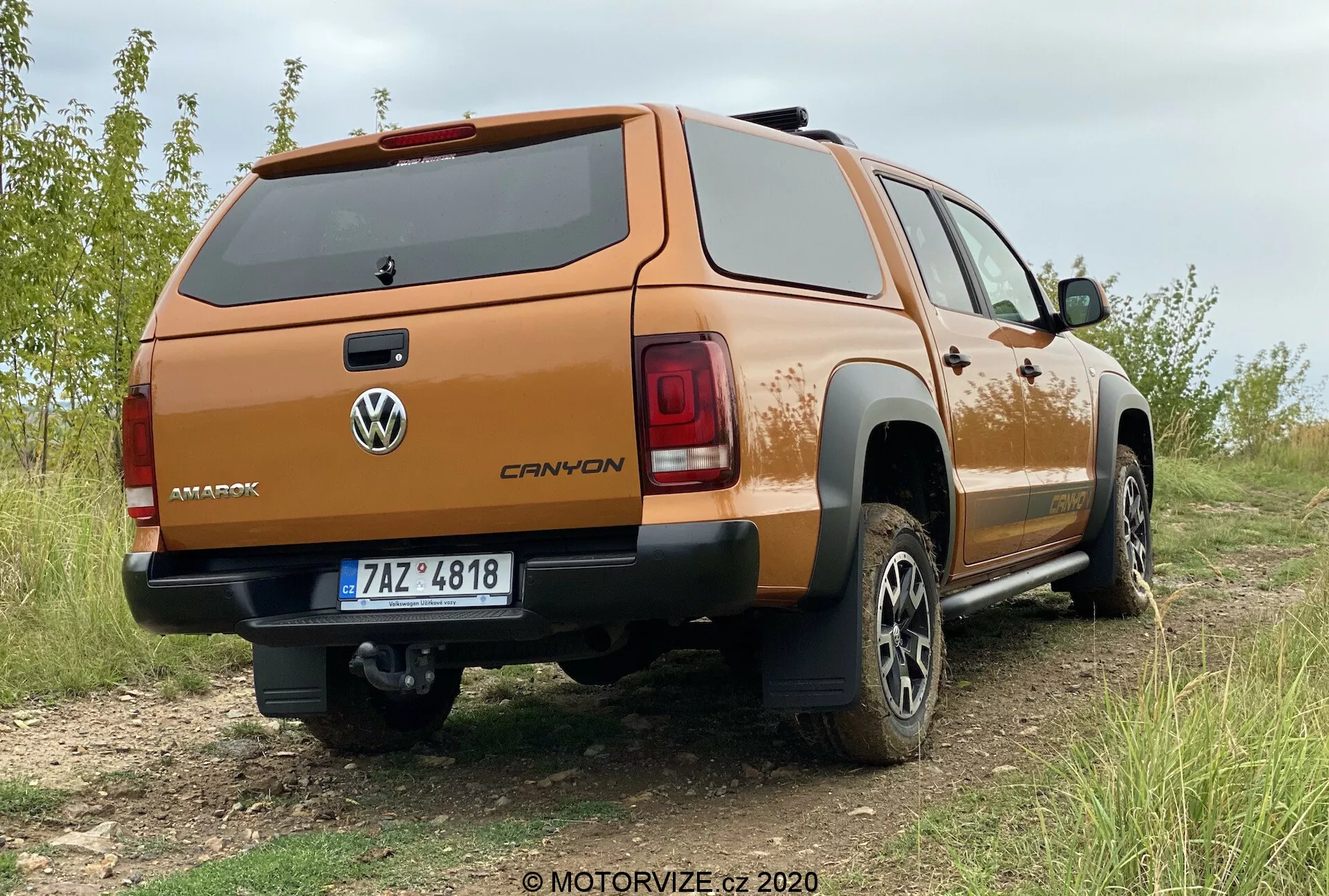 Uma vista profissional e detalhada de três quartos traseiros de um Volkswagen Amarok 2016, estacionado em um terreno levemente irregular e gramado. O lado traseiro direito do caminhão está em destaque, mostrando uma capota de carga fechada em um tom laranja ou cobre personalizado. A tampa traseira apresenta o logotipo da Volkswagen e o nome do modelo 'AMAROK'. As luzes traseiras têm design moderno e segmentado, e o para-choque traseiro inclui uma escada central, possivelmente equipada com sensores de estacionamento. A placa de licença tem uma moldura cromada ou polida e, sob o para-choque, é visível um engate de reboque, indicando capacidades de reboque. Os pneus todo-terreno e as rodas de liga de múltiplos raios sugerem prontidão off-road e um nível de acabamento superior. Uma proteção preta é visível nas laterais inferiores da cama do caminhão.