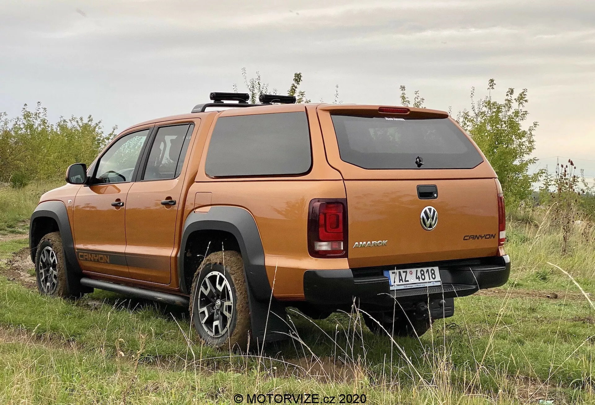 Vista traseira de três quartos de uma caminhonete Volkswagen Amarok 2016 laranja, mostrando o design das rodas, o perfil lateral, a janela traseira, as luzes traseiras, o robusto para-choques traseiro e a cobertura rígida da caçamba, estacionada em uma superfície não pavimentada com vegetação, destacando suas capacidades off-road.