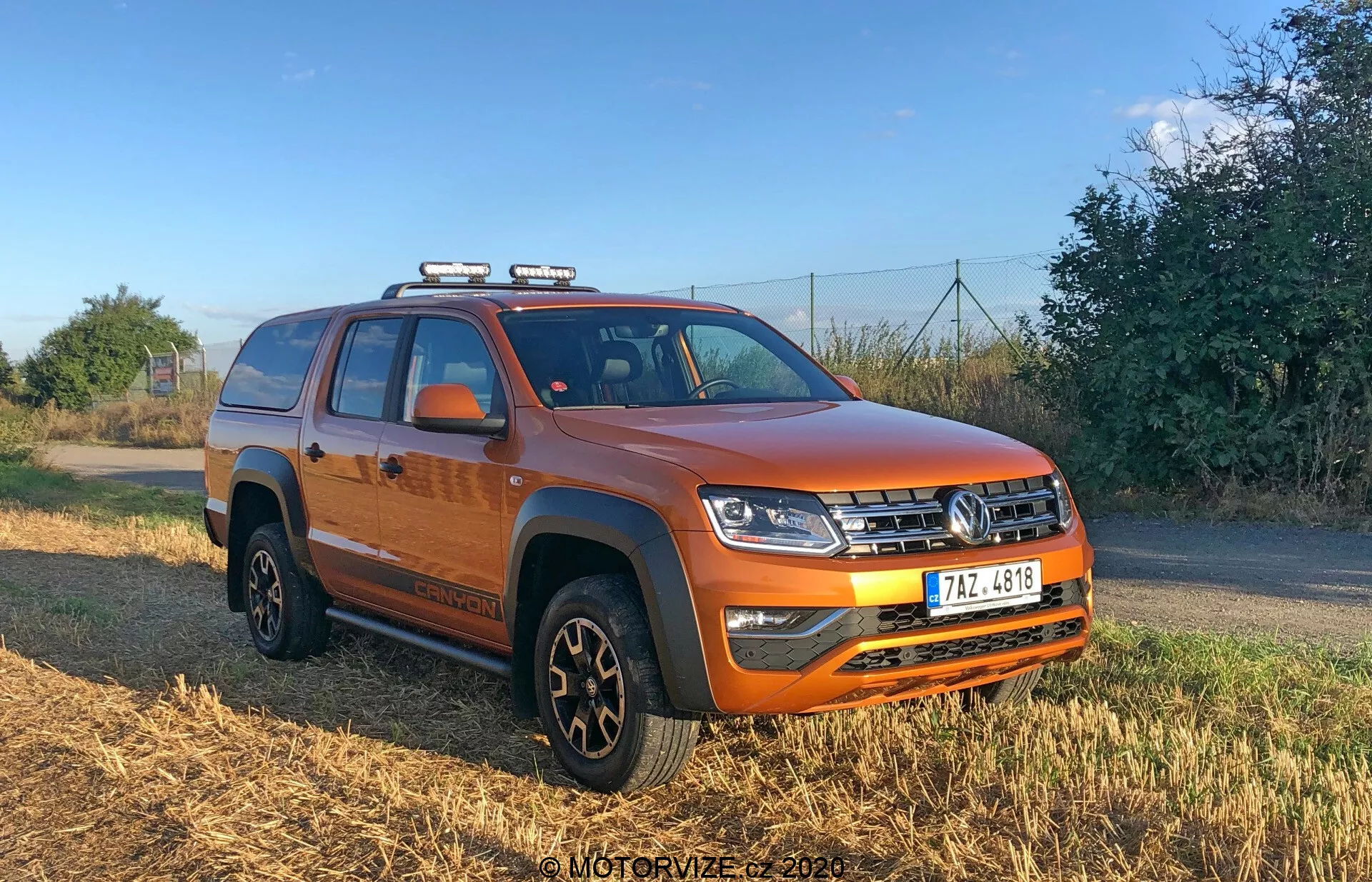 Imagem de uma vibrante caminhonete laranja VW Nutzfahrzeuge Amarok 2016 de quatro portas tirada em um ângulo de três quartos, mostrando a frente e o lado do veículo. A imagem destaca sua grade frontal com design horizontal, faróis de LED complexos, luzes auxiliares montadas no teto, para-choque robusto com faróis de neblina, placa de proteção, seção central elevada do capô, revestimento protetor preto nos arcos das rodas, rodas pretas com pneus multiuso, degraus laterais, espelhos com indicadores de direção integrados, baú coberto com uma lona e trilhos no teto para capacidade de armazenamento adicional.
