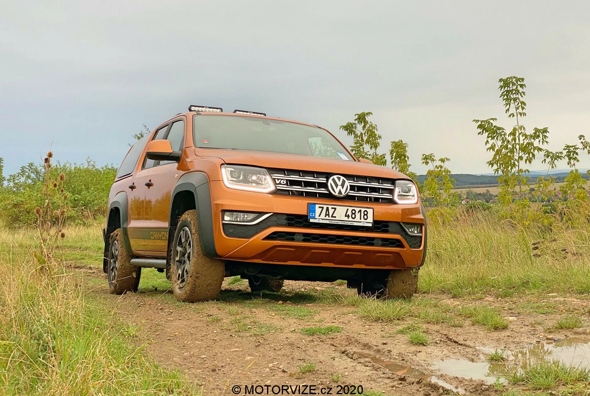 Ângulo baixo de três quartos dianteiro de um Volkswagen Amarok 2016 em um campo lamacento, exibindo sua pintura laranja, grade frontal com emblema Volkswagen, faróis, design das rodas e capacidade off-road. A robustez do veículo é destacada pela lama nos pneus e partes inferiores, com as rodas dianteiras viradas para a esquerda, enfatizando os padrões de rastro e altura de condução. Em um cenário de iluminação suave, seja de manhã ou no final da tarde, a imagem destaca a utilidade off-road do Amarok e seu avançado sistema de iluminação. A placa de licença é visível e deve ser obscurecida para privacidade.